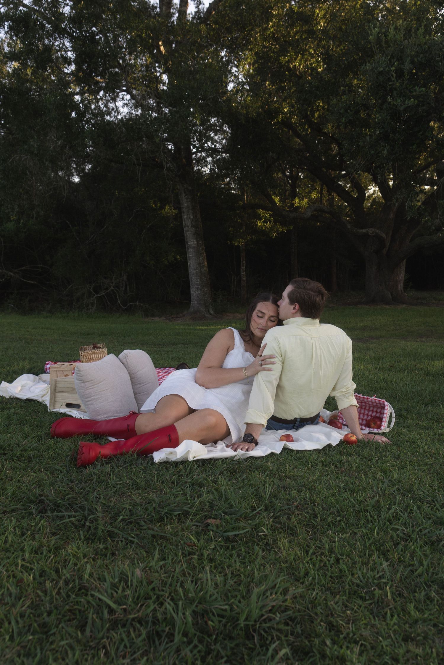 Couple sharing a romantic kiss during a picnic on a blanket in a lush green park surrounded by trees.