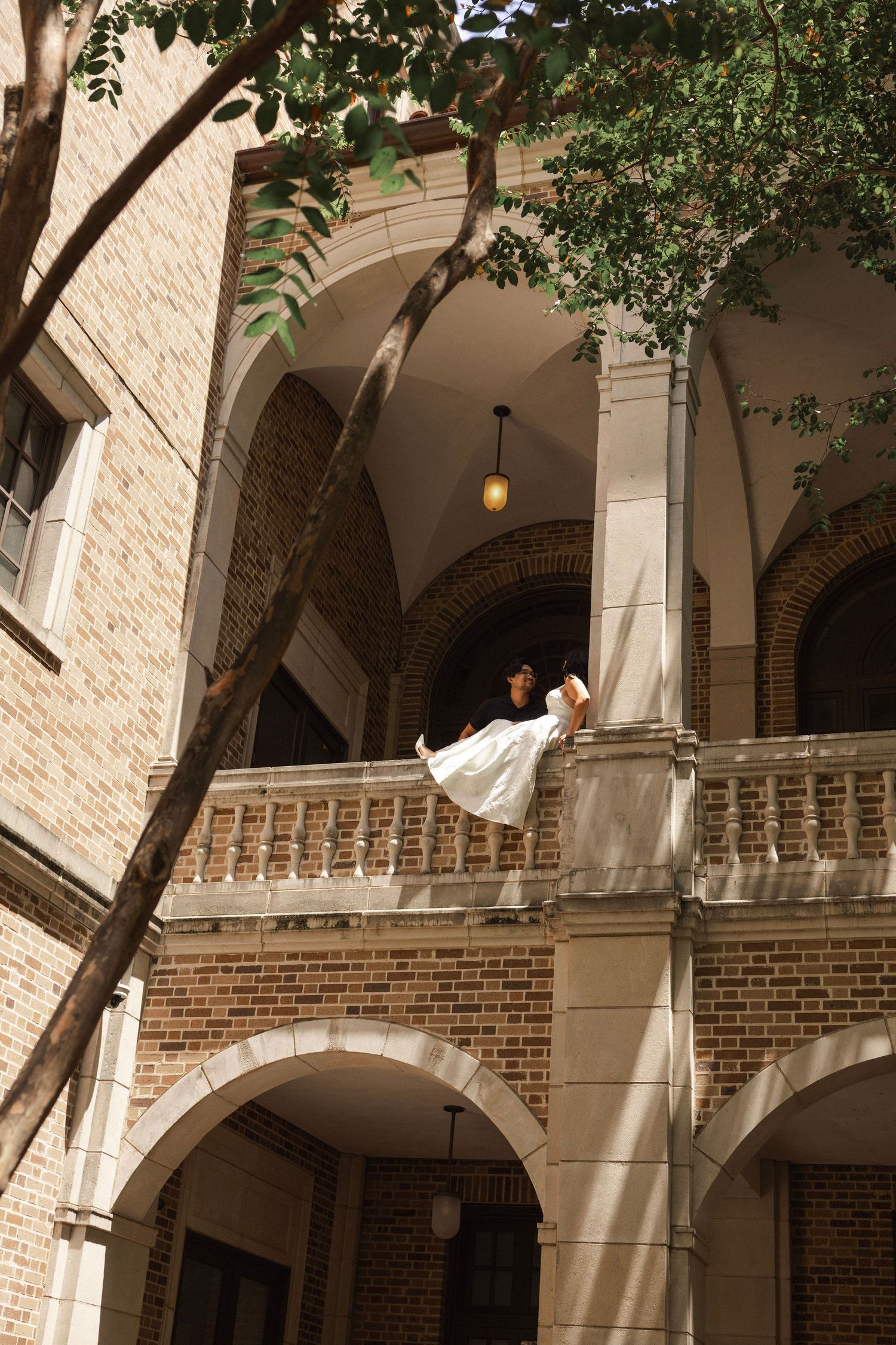 Woman in white dress poses on ornate brick building balcony with arched colonnade and hanging lantern.