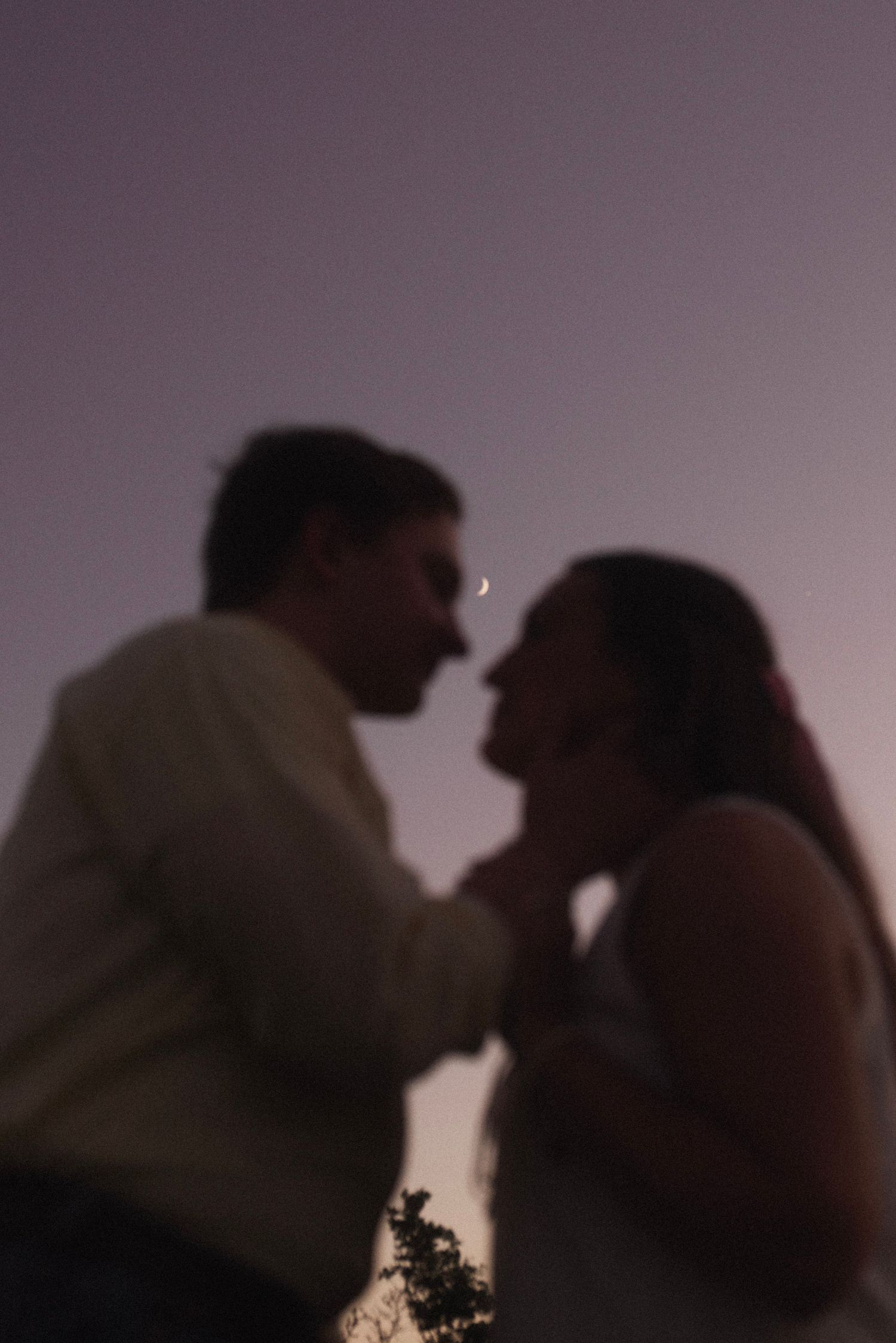 Romantic couple silhouetted against a twilight purple sky with a crescent moon, sharing an intimate moment at dusk.