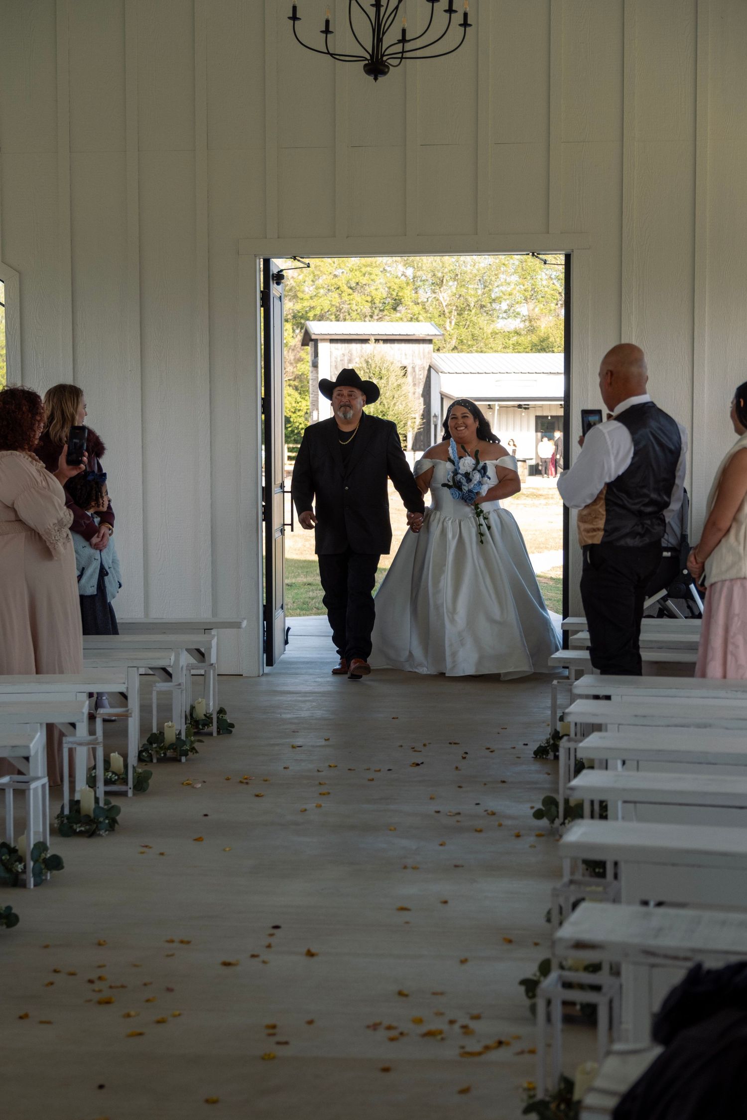 Bride in white ballgown walking down aisle escorted by man in black cowboy hat in white chapel.