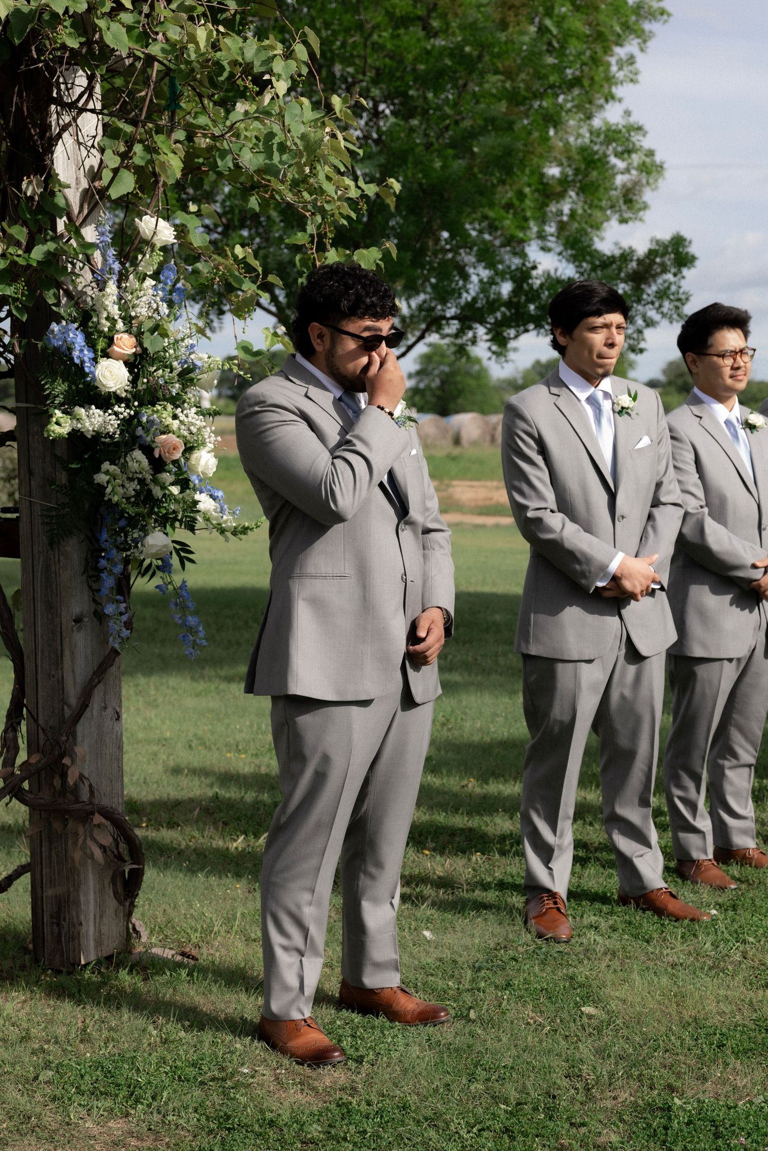 Emotional groom crying in gray suit at outdoor wedding ceremony beside floral arch with groomsmen standing behind.