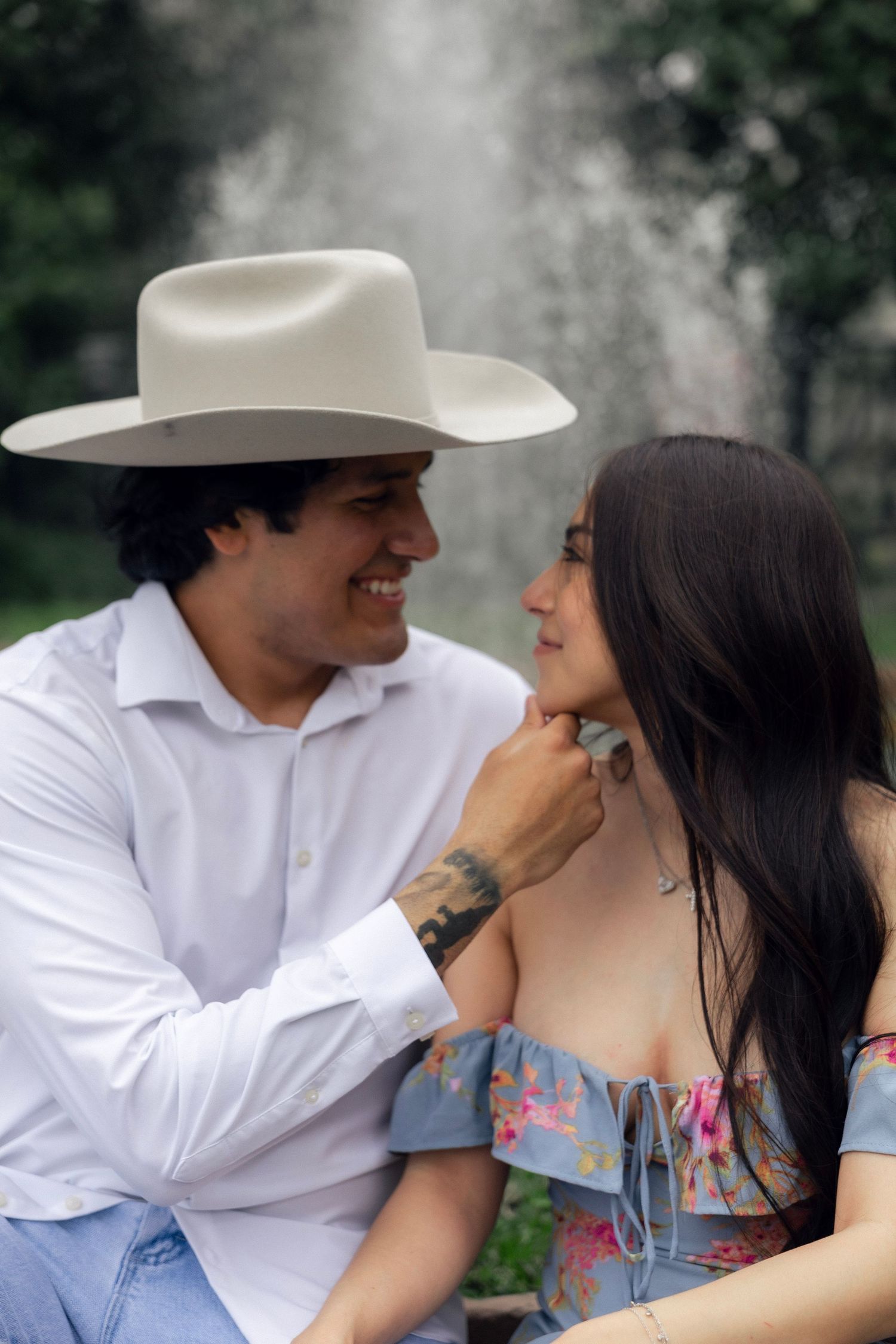 Smiling man in white shirt and wide-brim hat gently touches womans chin during romantic outdoor moment.