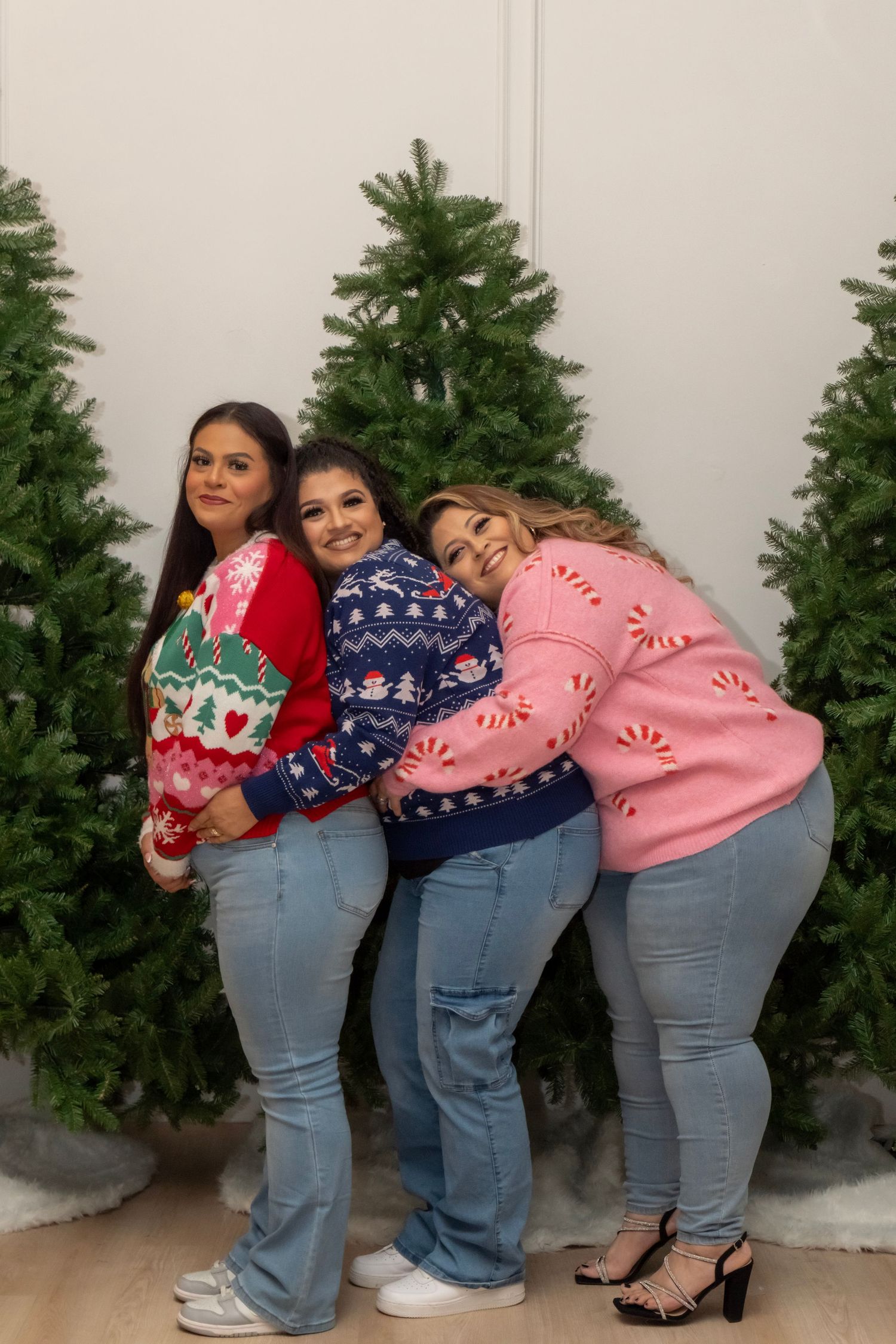 Three women in festive Christmas sweaters and jeans pose together in front of green Christmas trees.