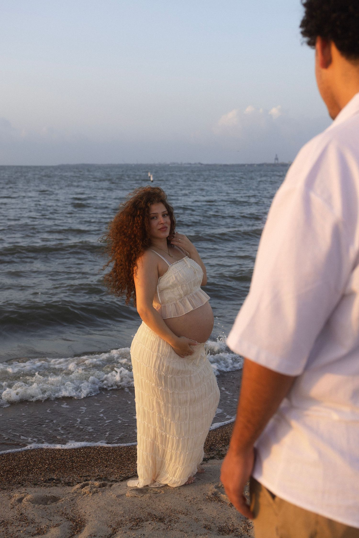 Pregnant woman in white dress stands on sandy beach at sunset, cradling her belly while a man in white shirt looks on.