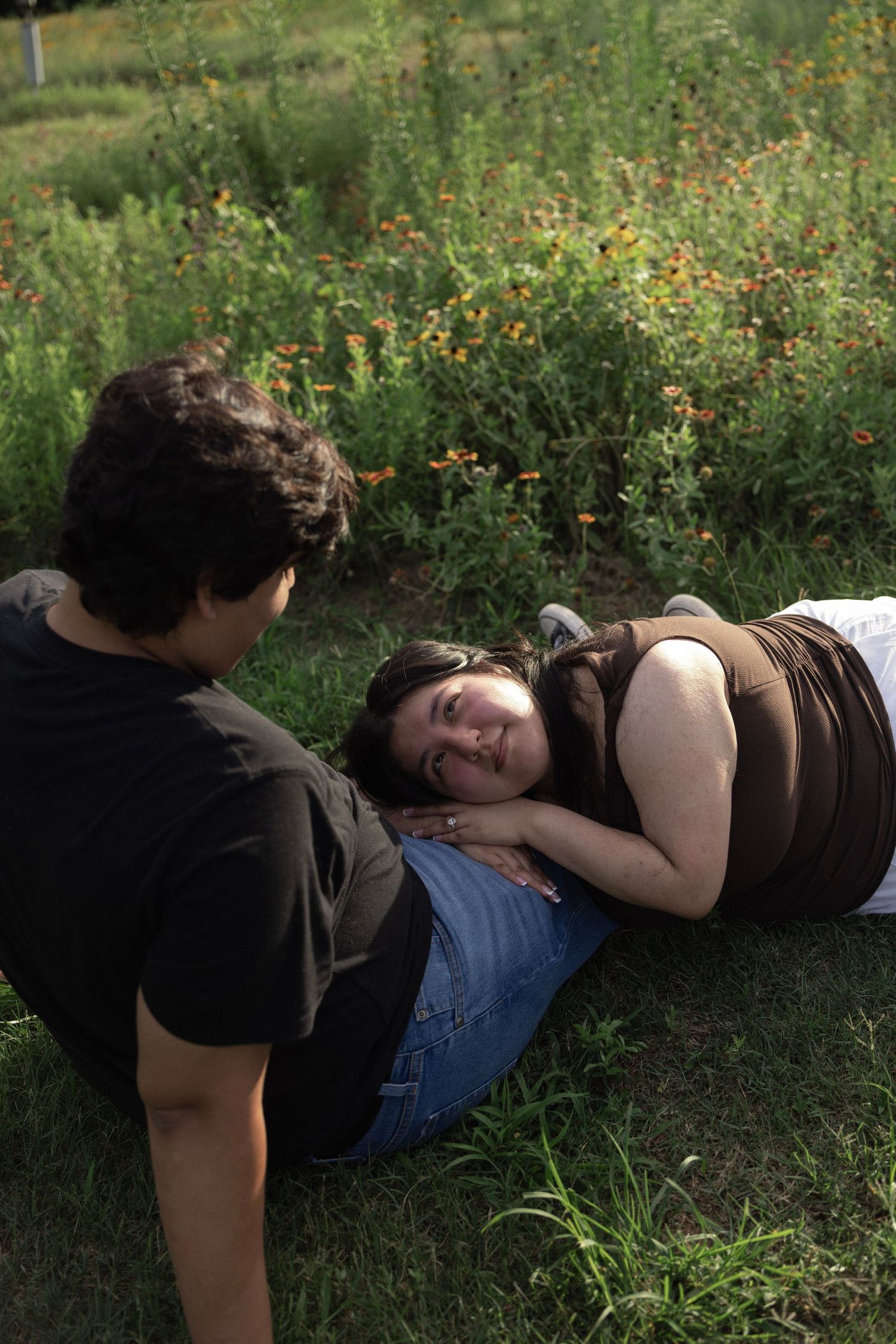 A young woman rests her head on a man's lap while lying in green grass near a garden of wildflowers.