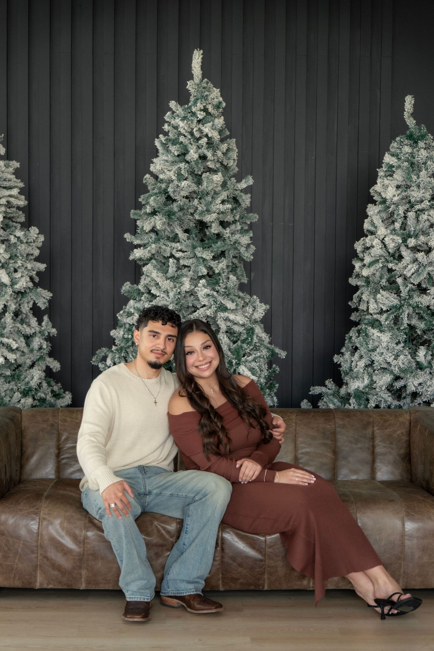 A couple sits on a leather sofa in front of snow-flocked Christmas trees, smiling warmly in a festive holiday portrait.