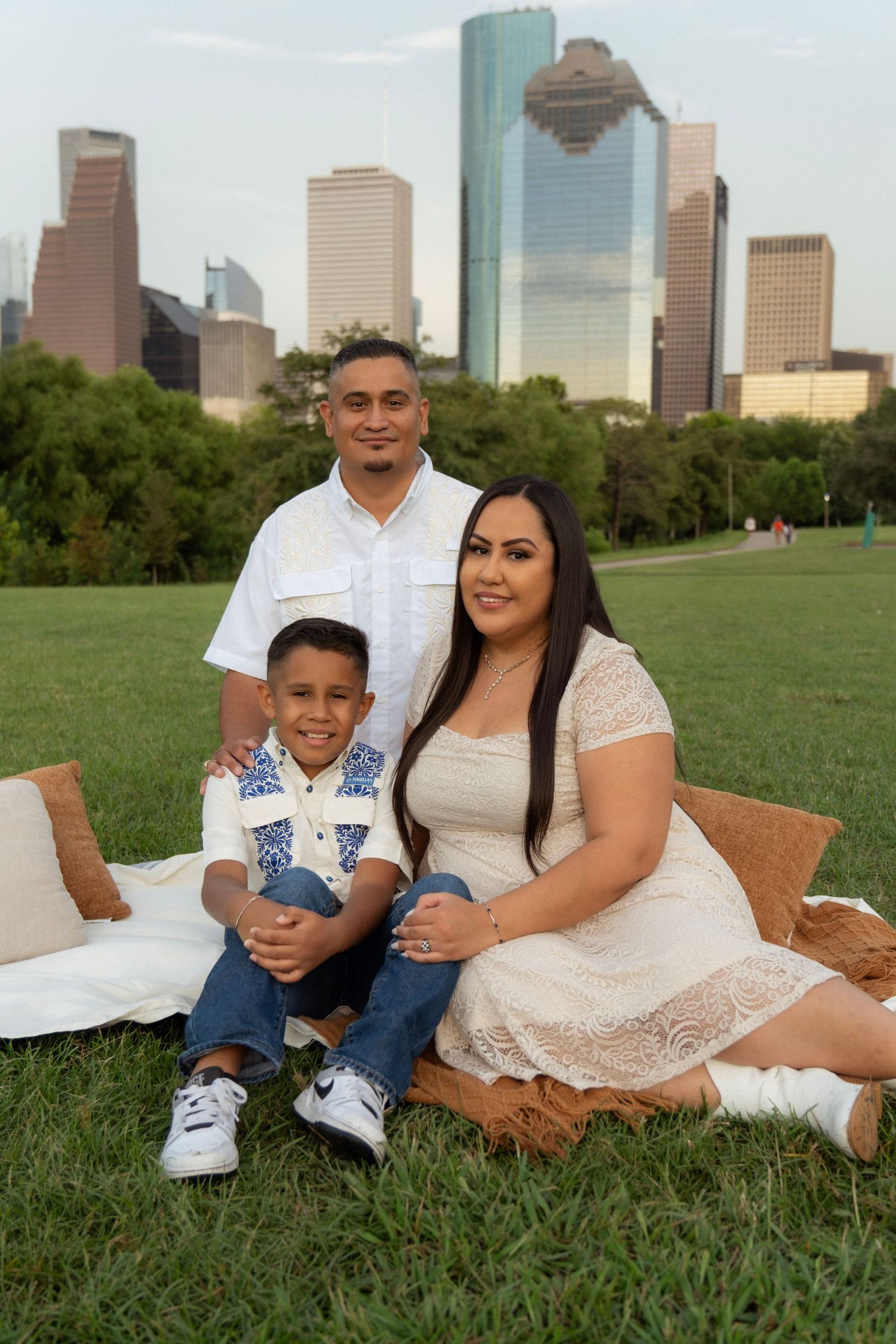 Family of three posing on grass with Houston skyline in background, wearing white and blue outfits during golden hour.