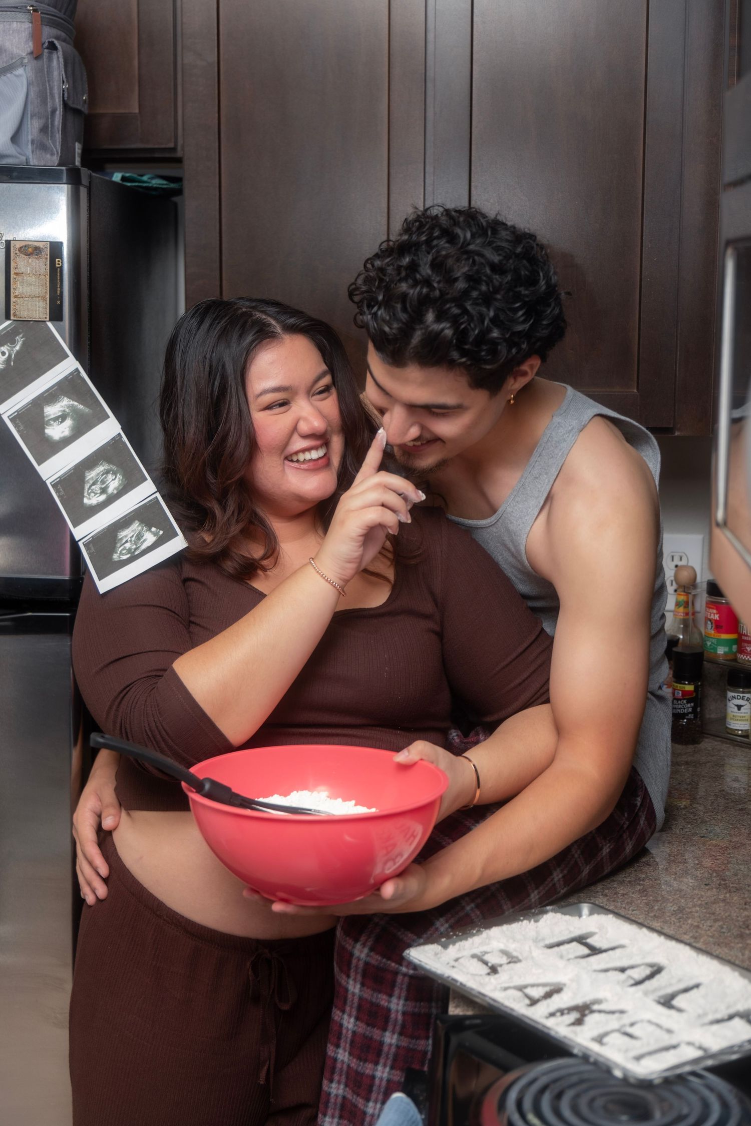 Laughing pregnant couple in kitchen with ultrasound photos and half baked pregnancy announcement.
