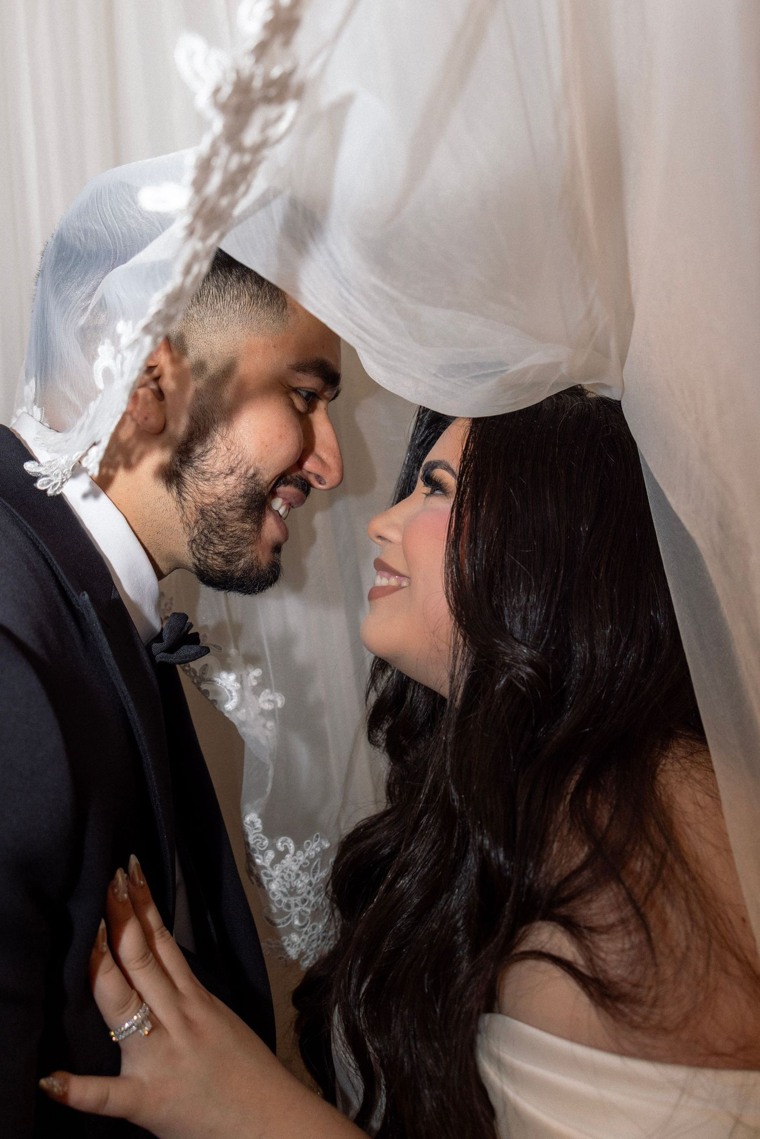 Romantic bride and groom smiling at each other under flowing white lace wedding veil.