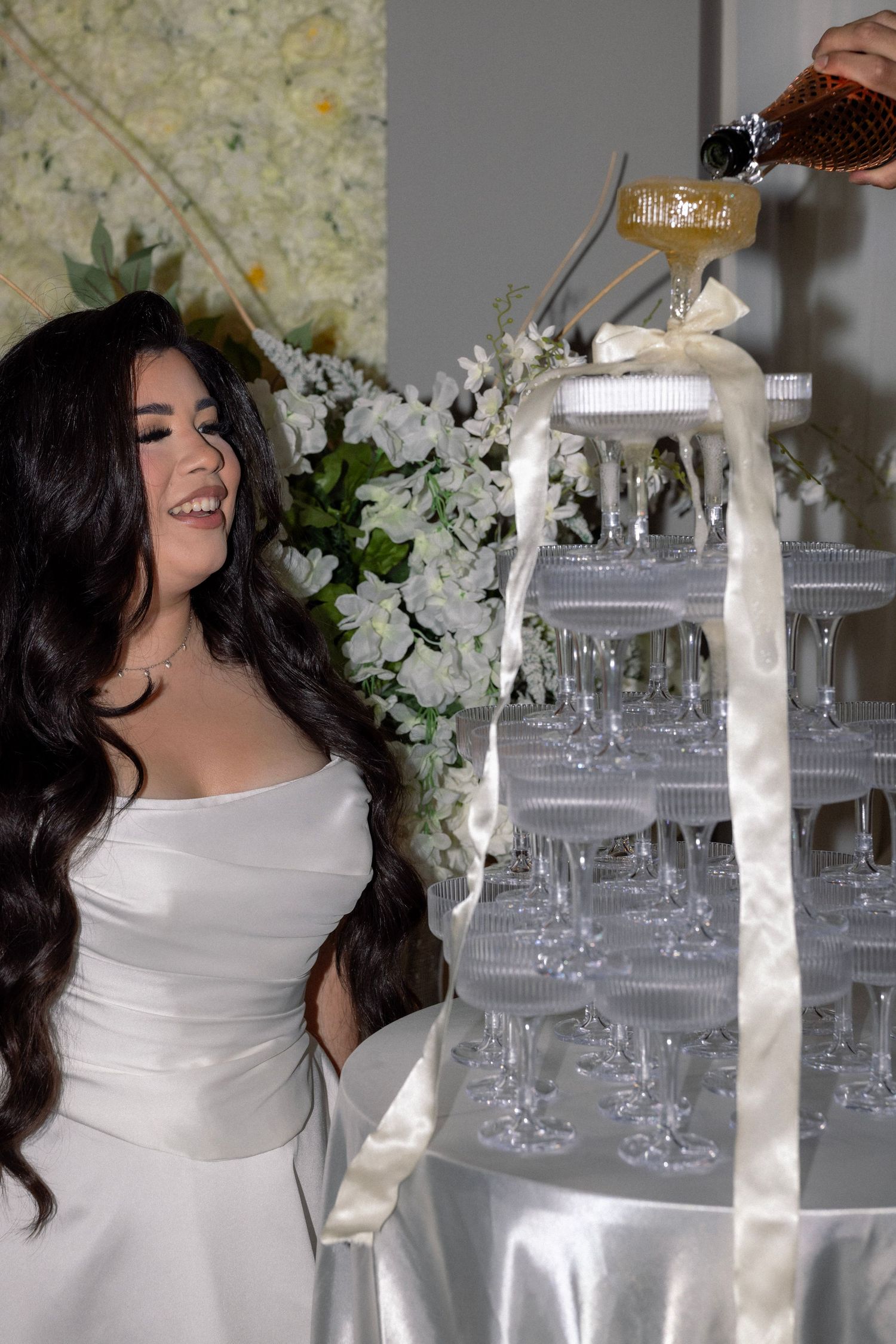 Smiling bride in white strapless gown watches champagne tower being filled at wedding reception.
