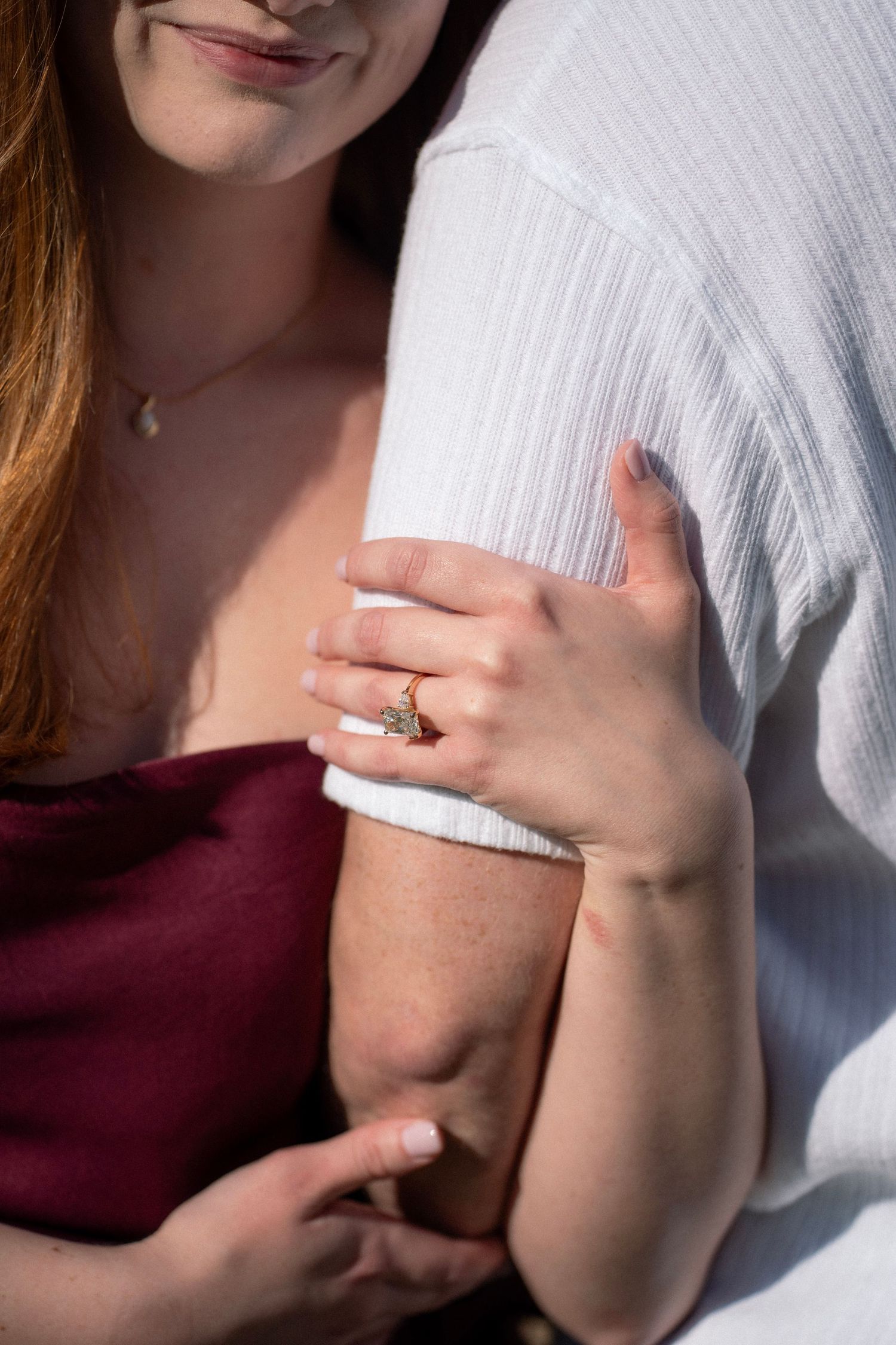 Close-up of woman in burgundy dress showing engagement ring while embracing partner in white.