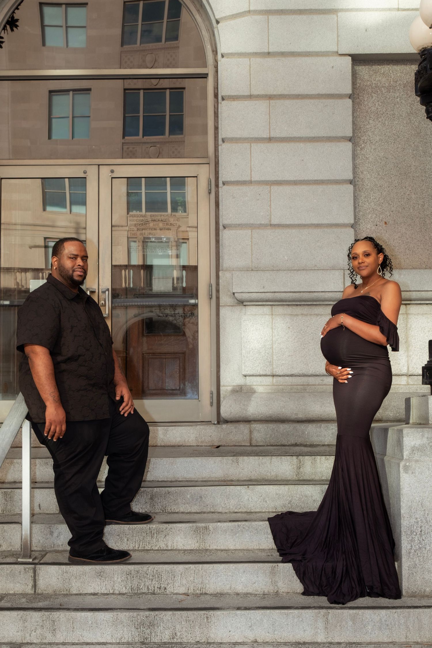 Pregnant couple posing on stone steps, woman in elegant black gown cradling baby bump.