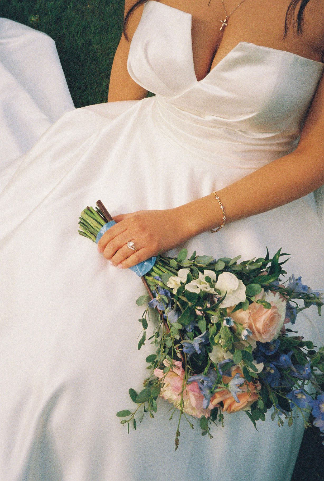 Bride in white satin strapless gown holding lush bouquet of pink, blue, and white flowers with greenery outdoors.