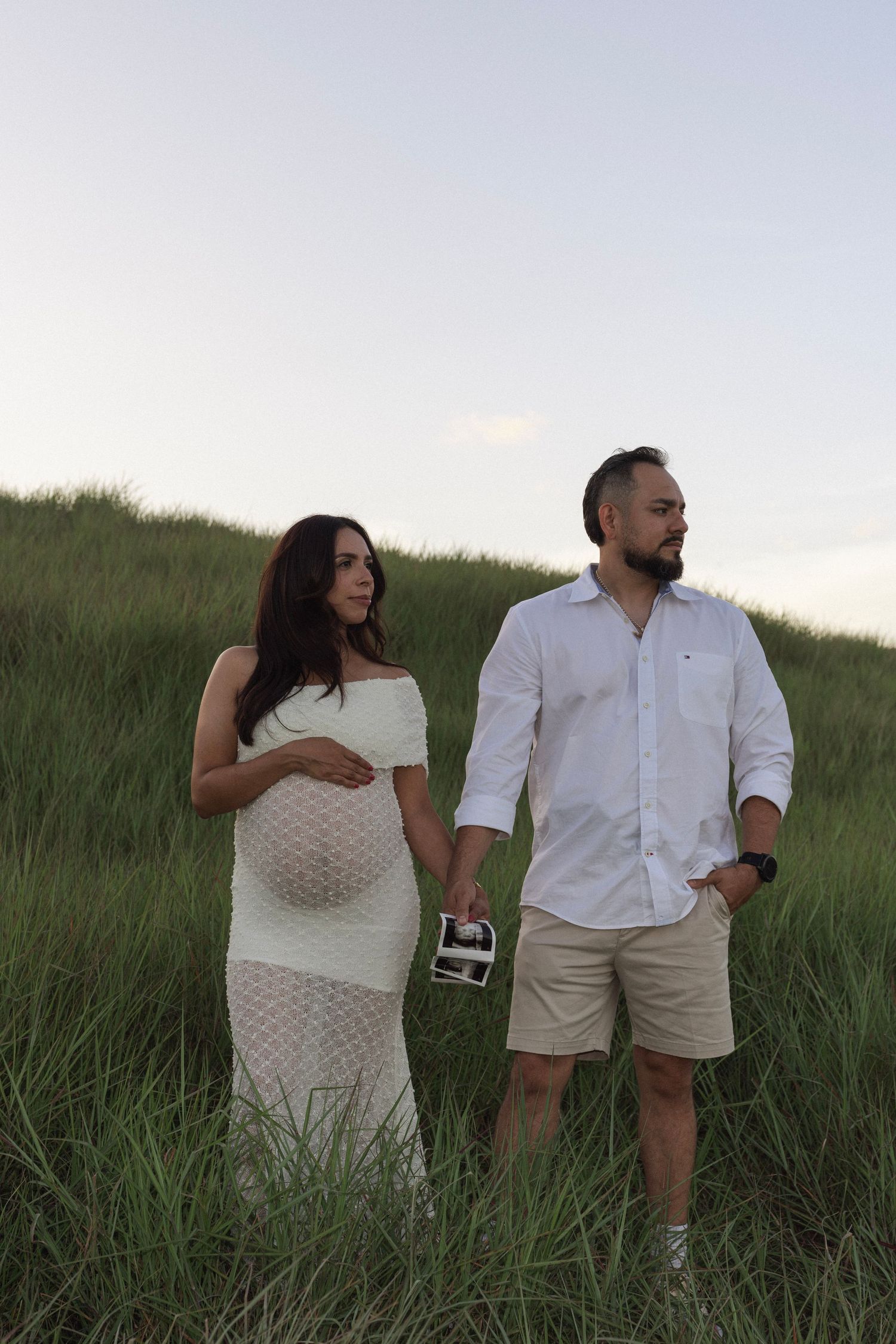 Pregnant couple standing in green grass field, woman in white lace dress holding belly, man in white shirt holding ultrasound photo.