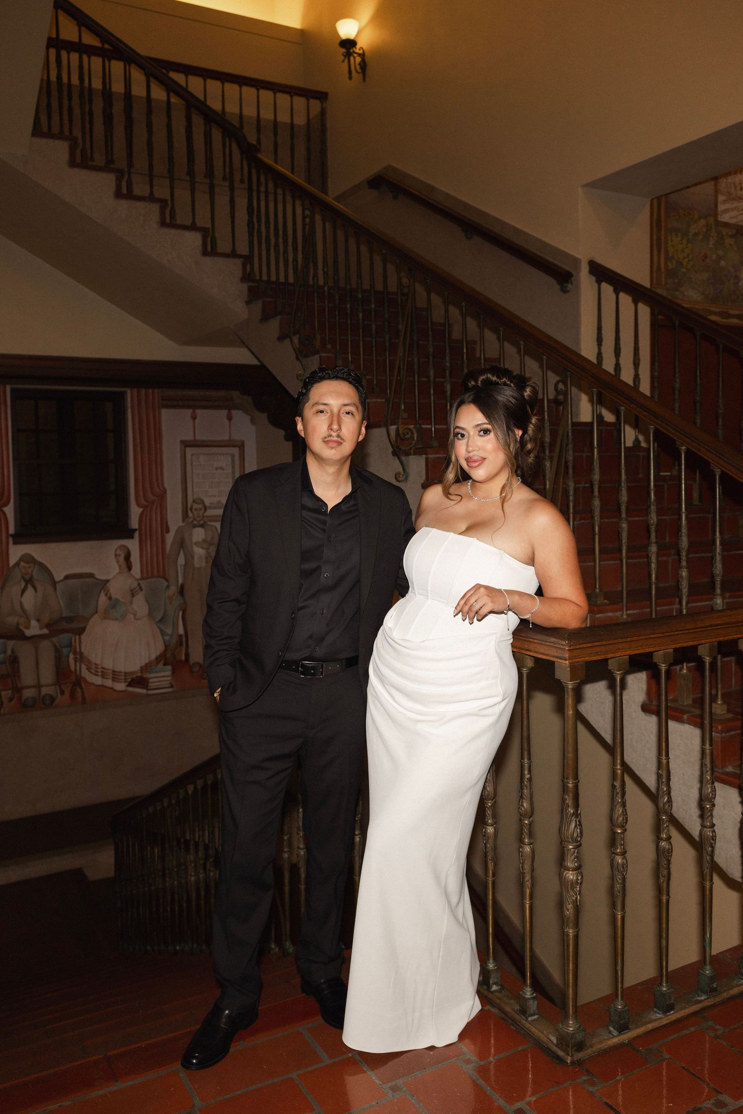 Elegant couple posing by ornate wooden staircase, woman in white strapless gown and man in black suit.