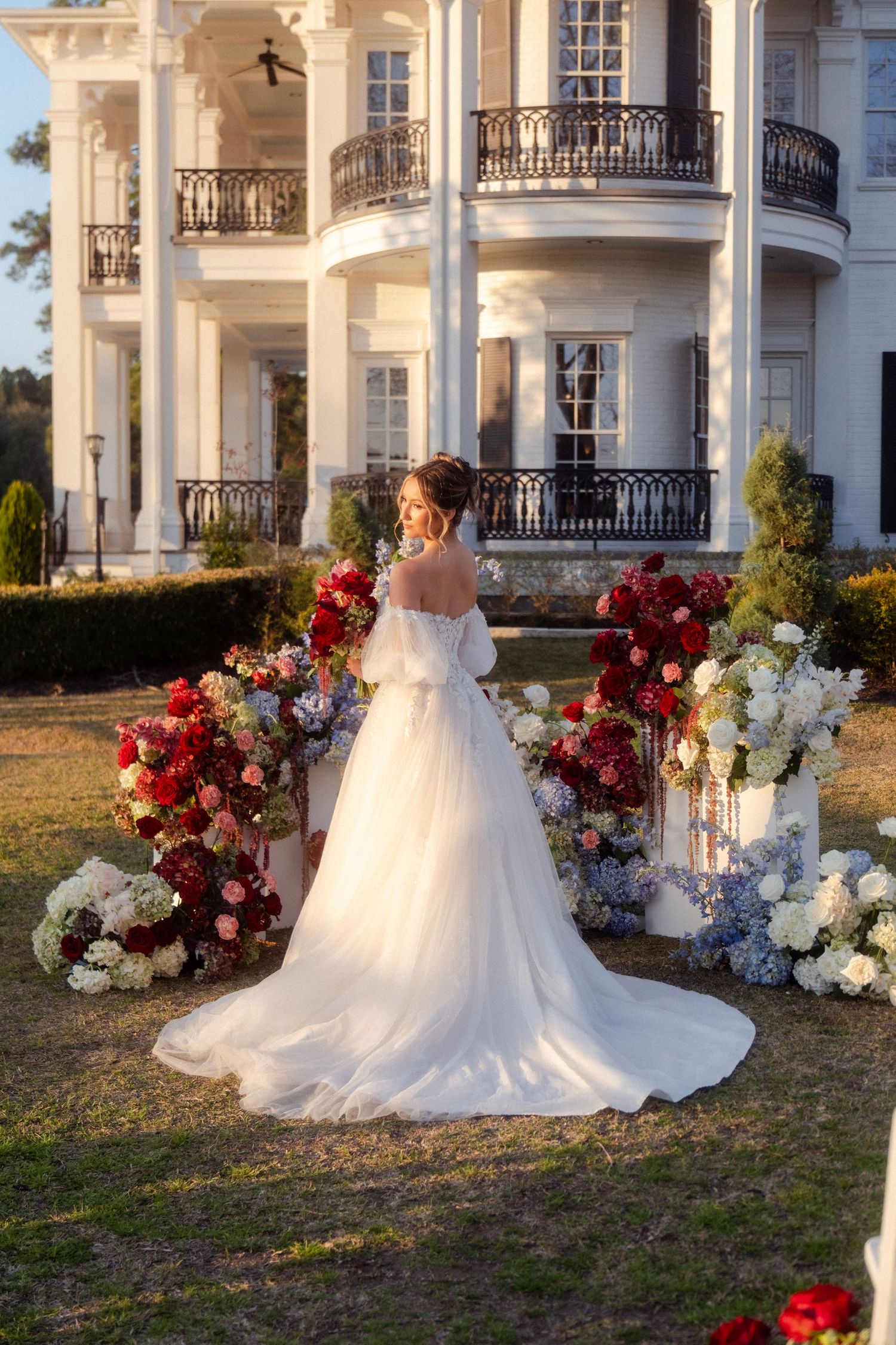 Bride in white off-shoulder gown stands before floral arch with red and white blooms at elegant white mansion.