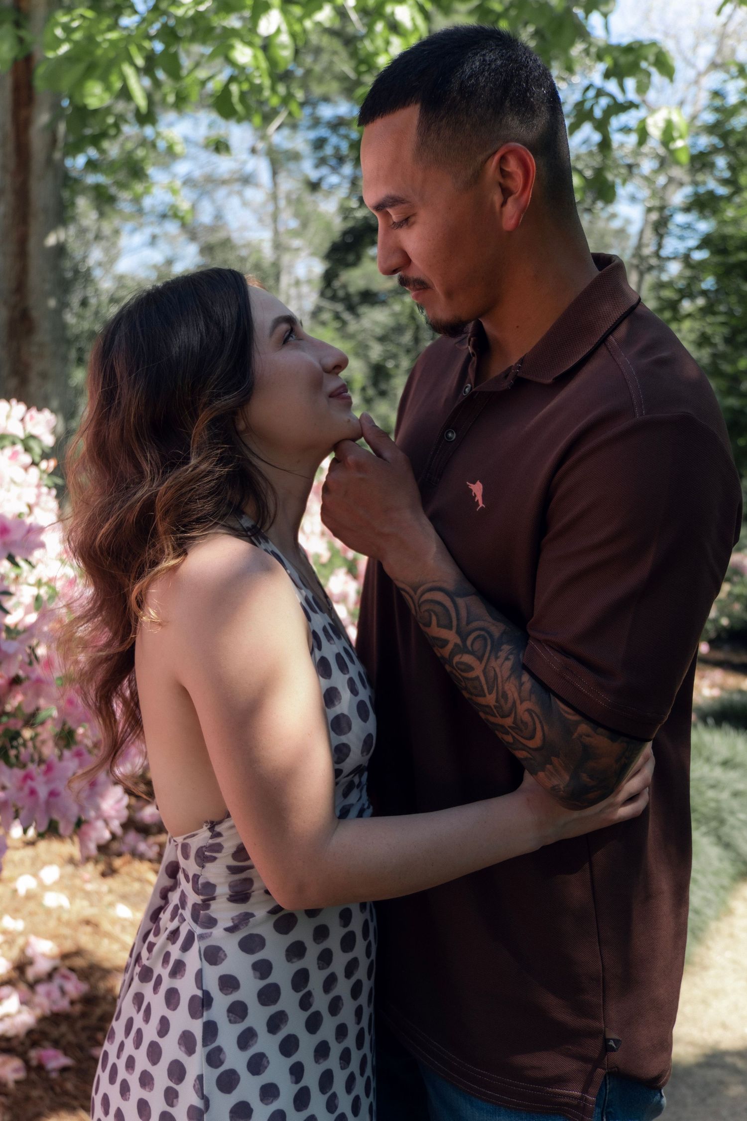 A couple shares a romantic moment outdoors, surrounded by pink flowers, as the man gently touches the woman's chin.