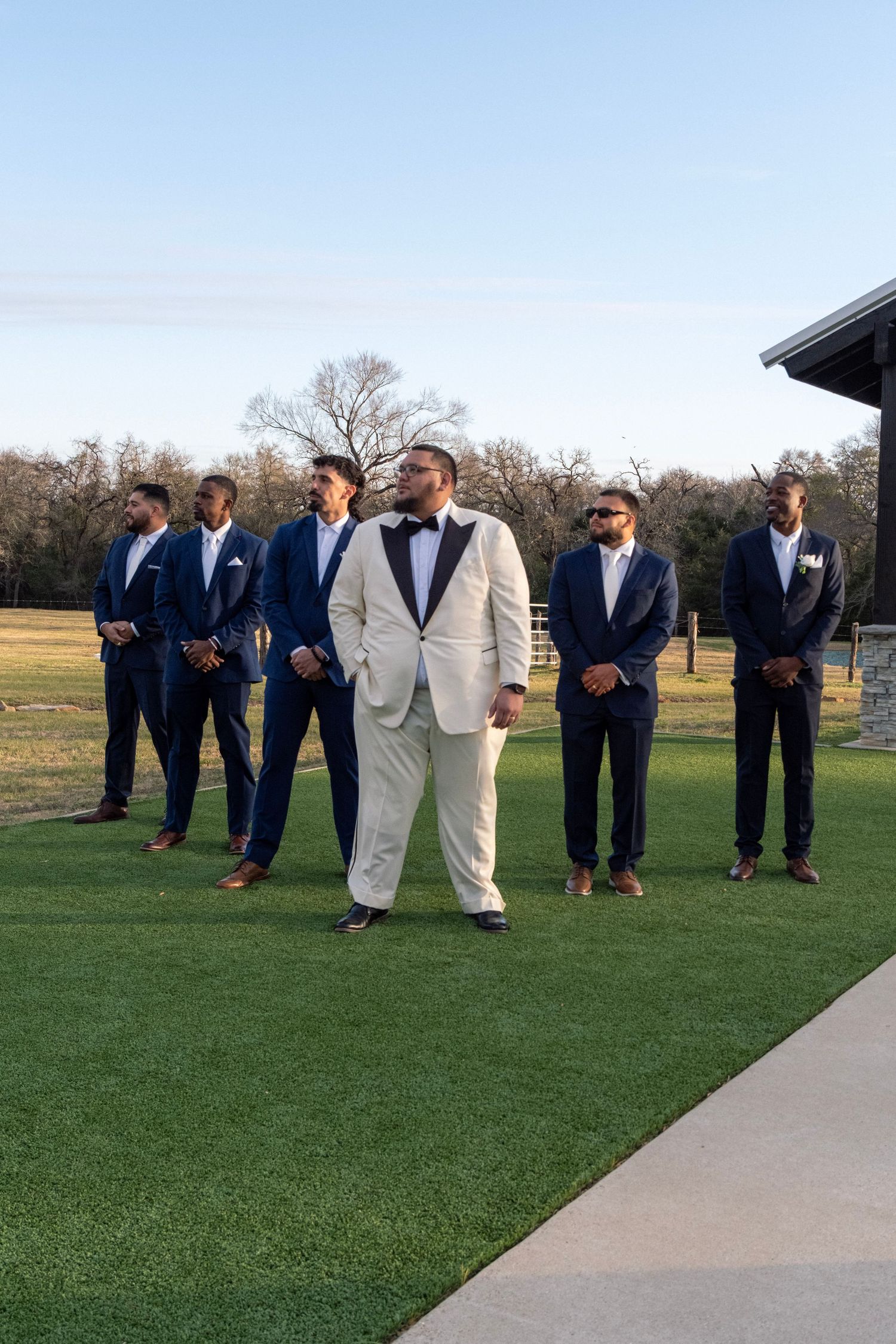 Groom in white tuxedo stands with groomsmen in navy suits on green lawn at outdoor wedding.