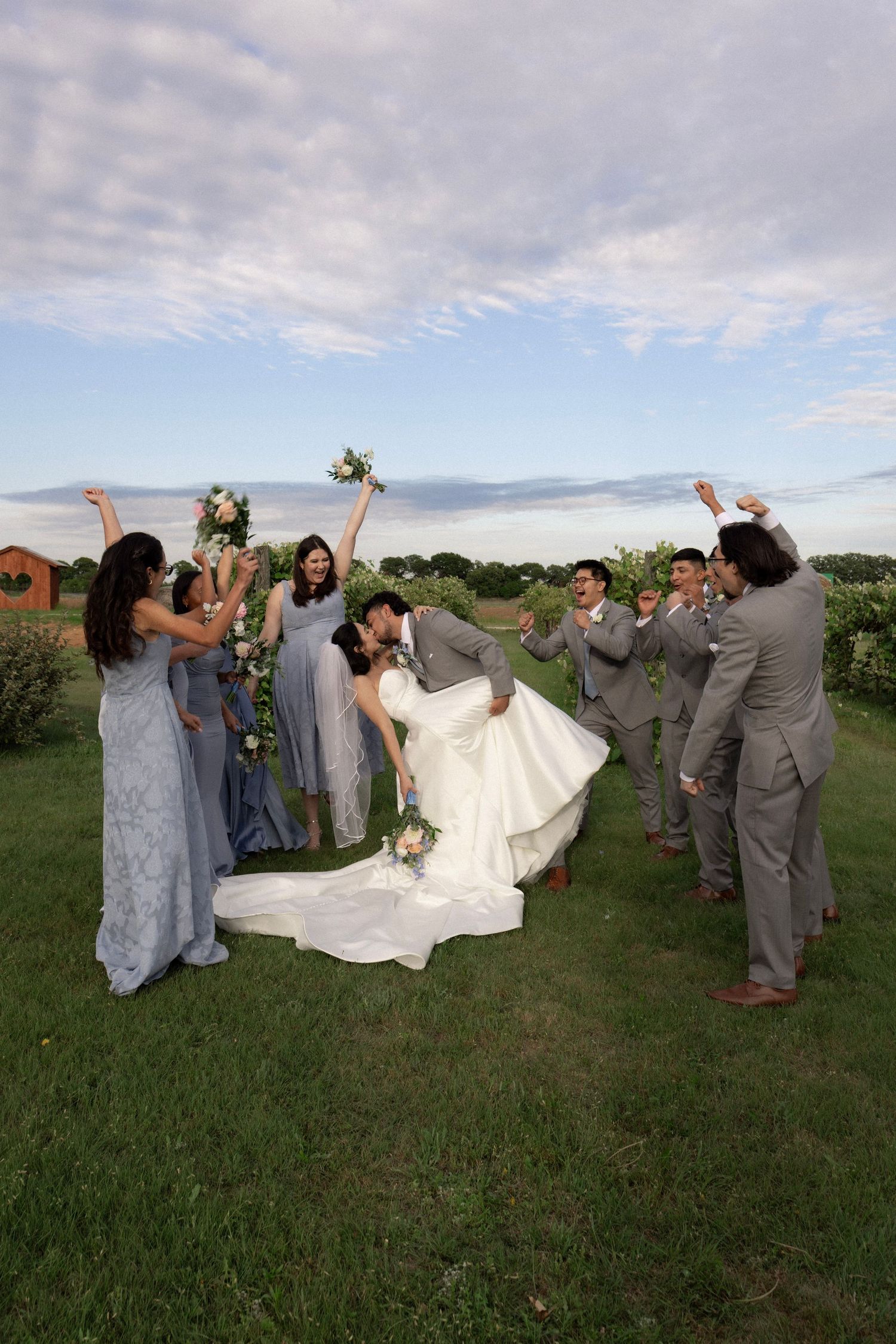 Wedding party celebrating as groom dips bride for kiss on green lawn with bridesmaids and groomsmen cheering.