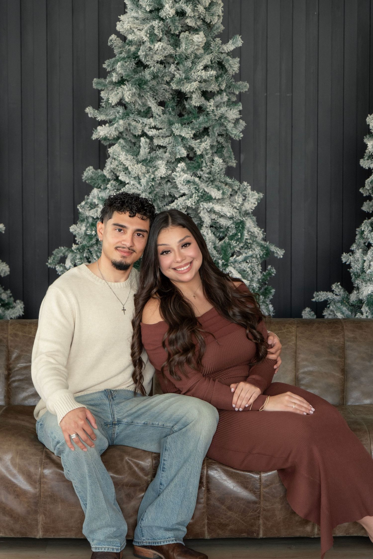 Couple sitting on leather couch in front of flocked Christmas trees, woman in brown dress, man in cream sweater and jeans.