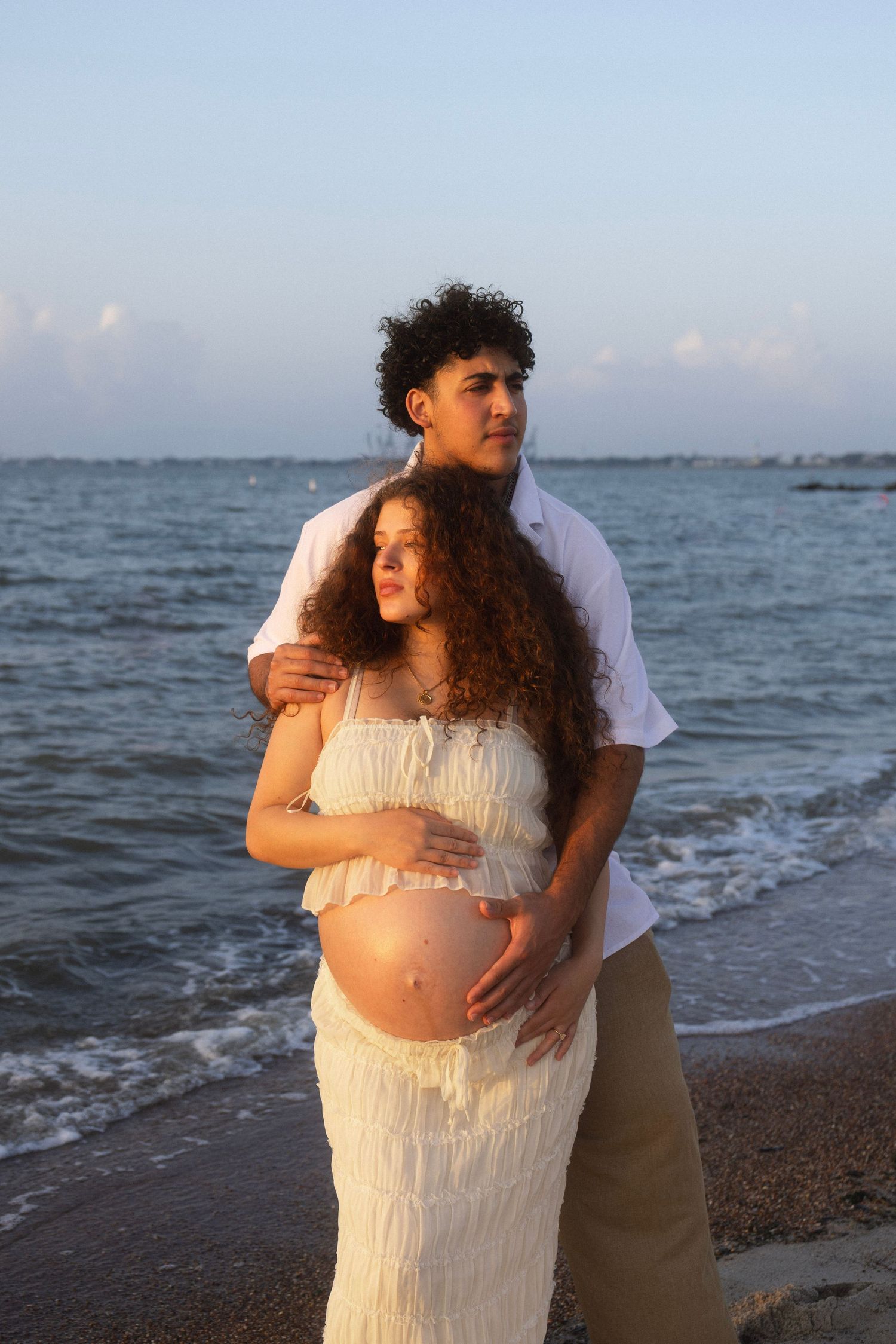Pregnant couple embracing on beach at sunset, woman in white dress cradling baby bump, man in white shirt behind her.