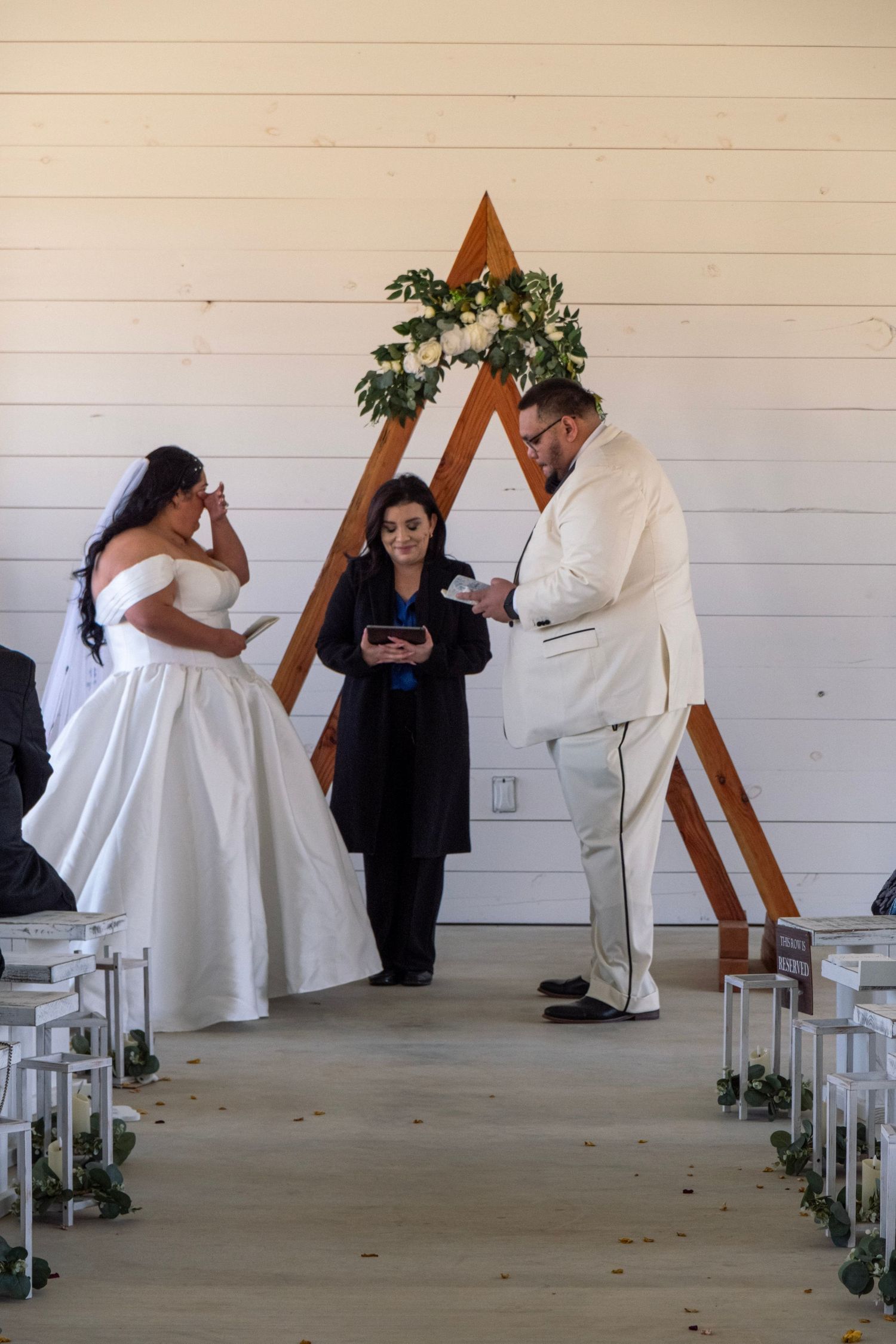 Bride and groom exchanging vows before officiant at modern triangular wooden arch in white chapel.