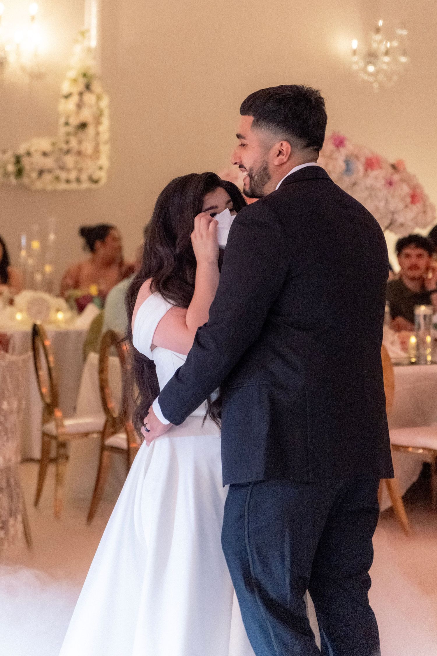 Bride and groom share emotional first dance in elegant reception hall with chandelier lighting.