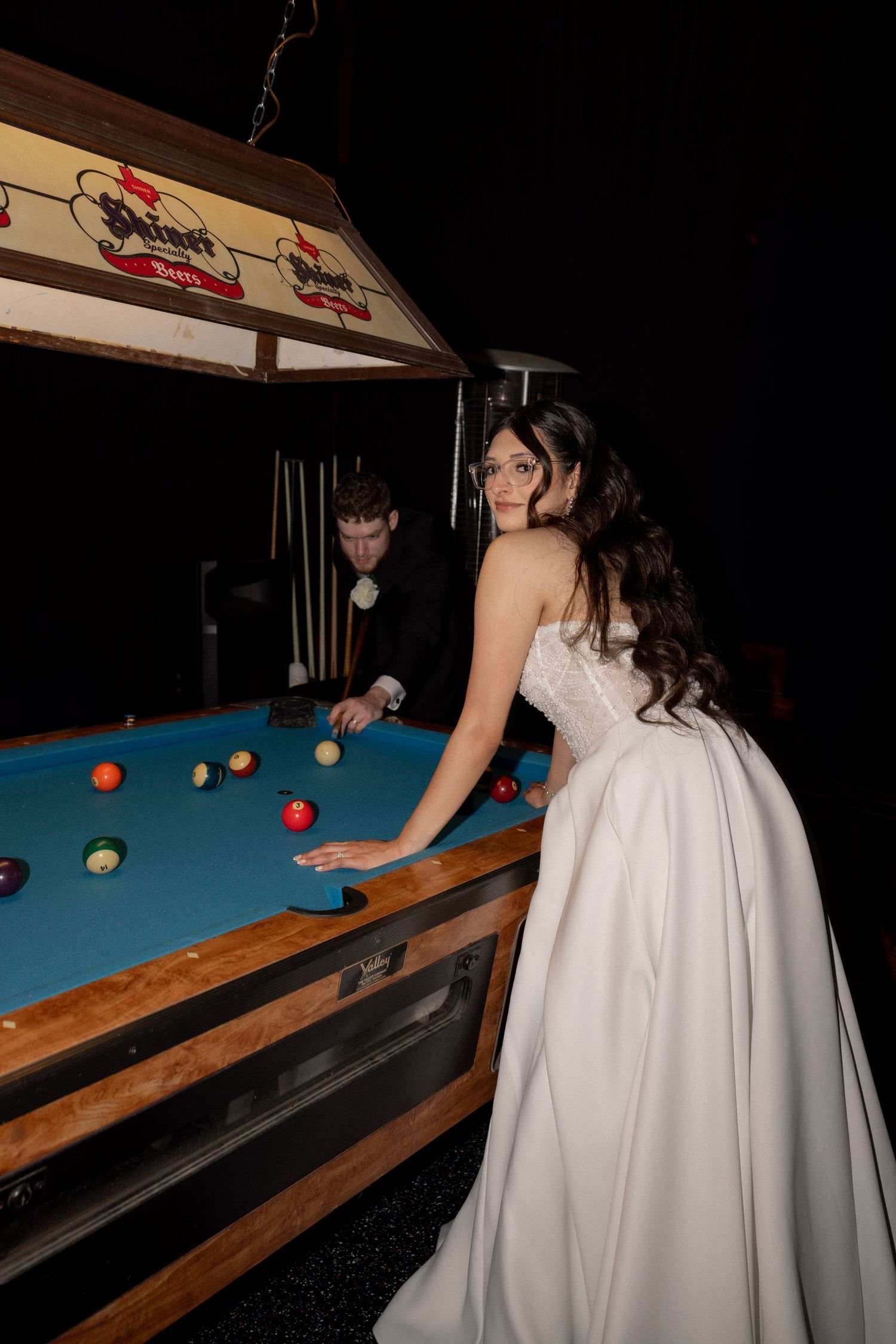 Bride in strapless white gown playing pool with groom at billiard table during wedding reception.
