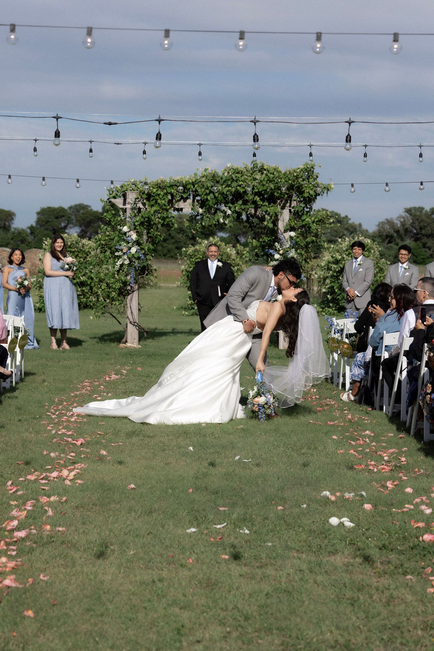Bride and groom sharing romantic dip kiss at outdoor wedding ceremony with petal-lined aisle and string lights.