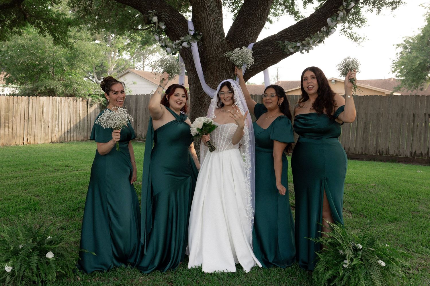 A bride in a white gown celebrates with four bridesmaids in emerald green dresses outdoors.