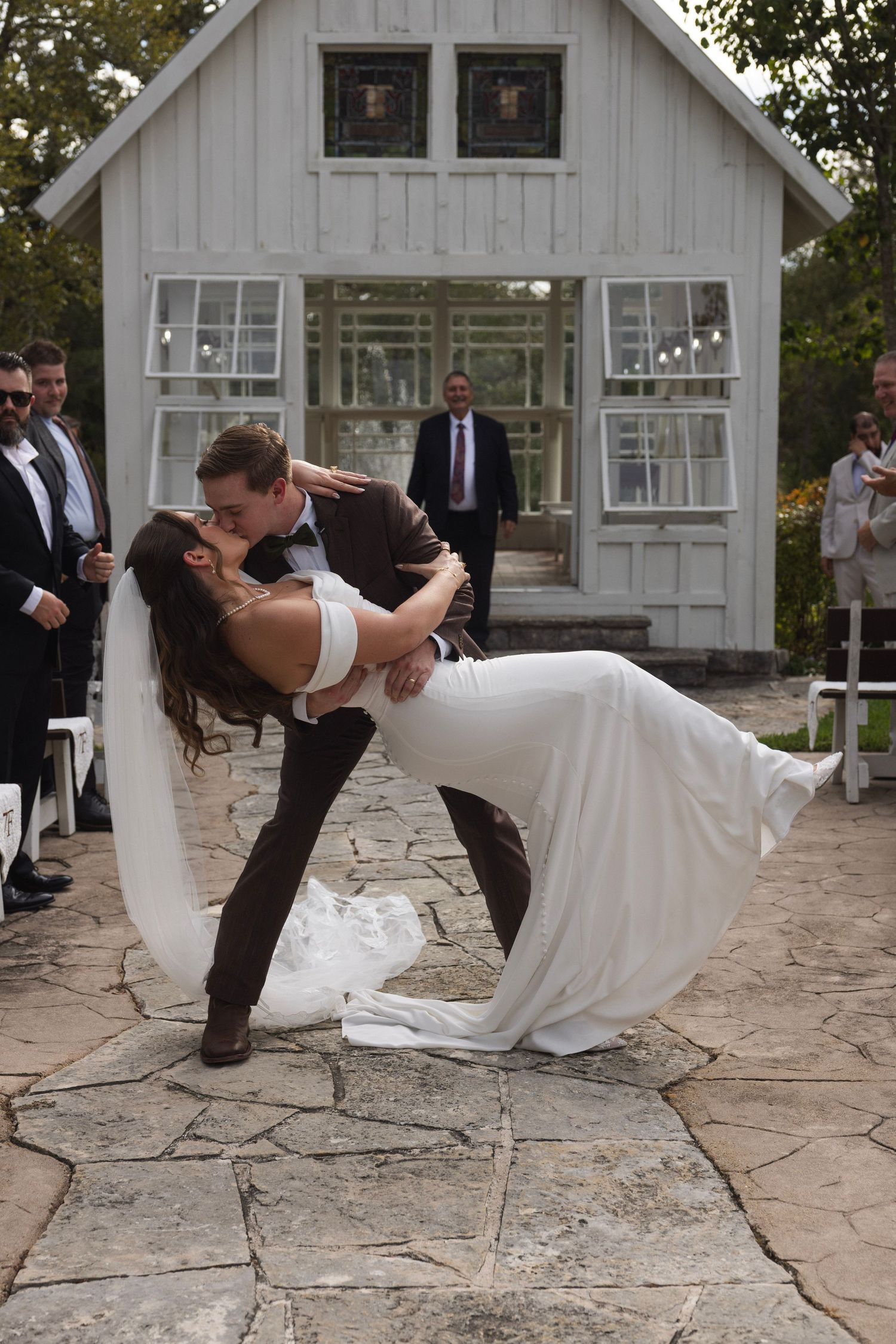 Groom dips bride for a romantic kiss outdoors at a rustic wedding venue with white chapel backdrop.