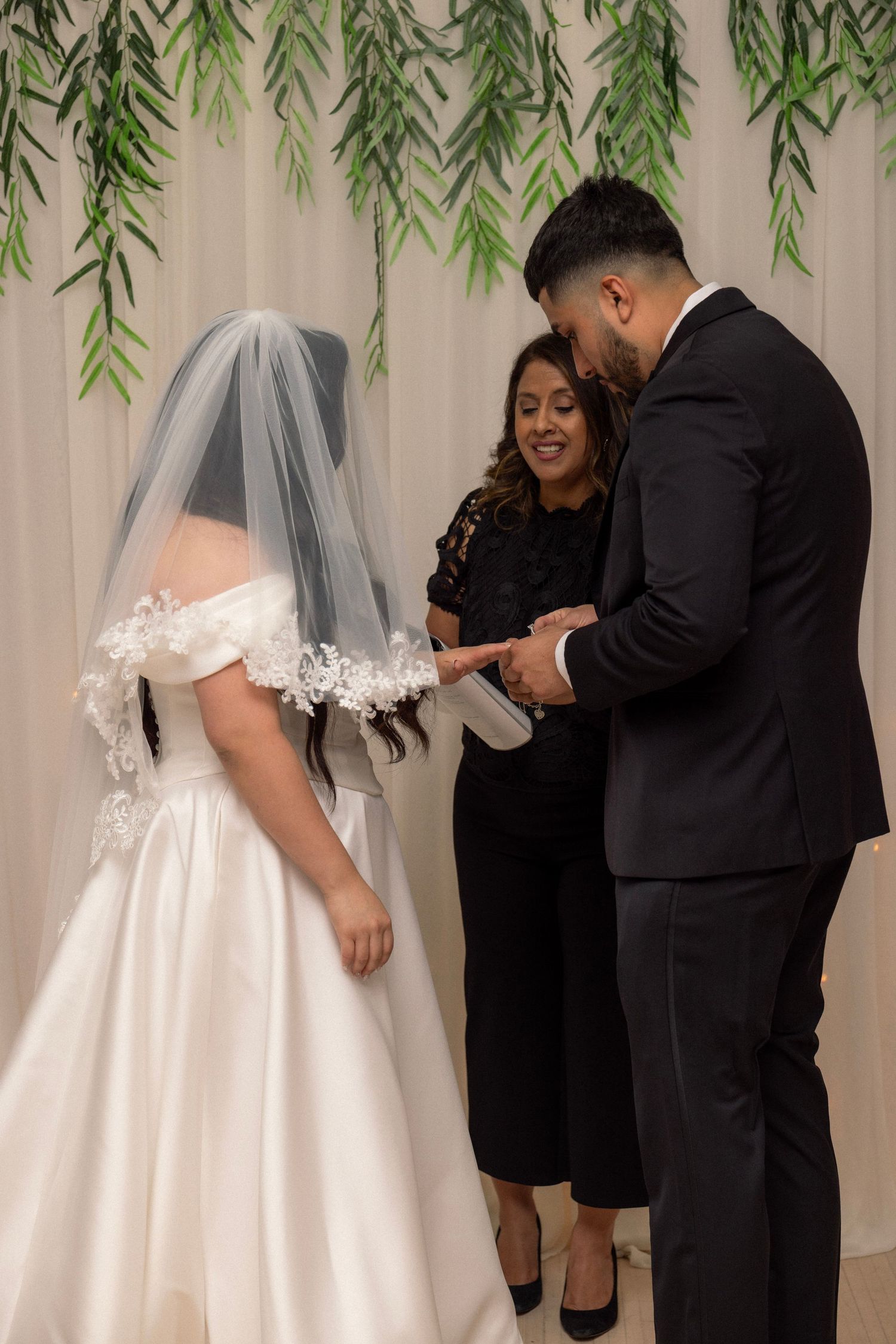 Bride and groom exchanging rings at wedding altar with greenery backdrop, officiant smiling behind them.