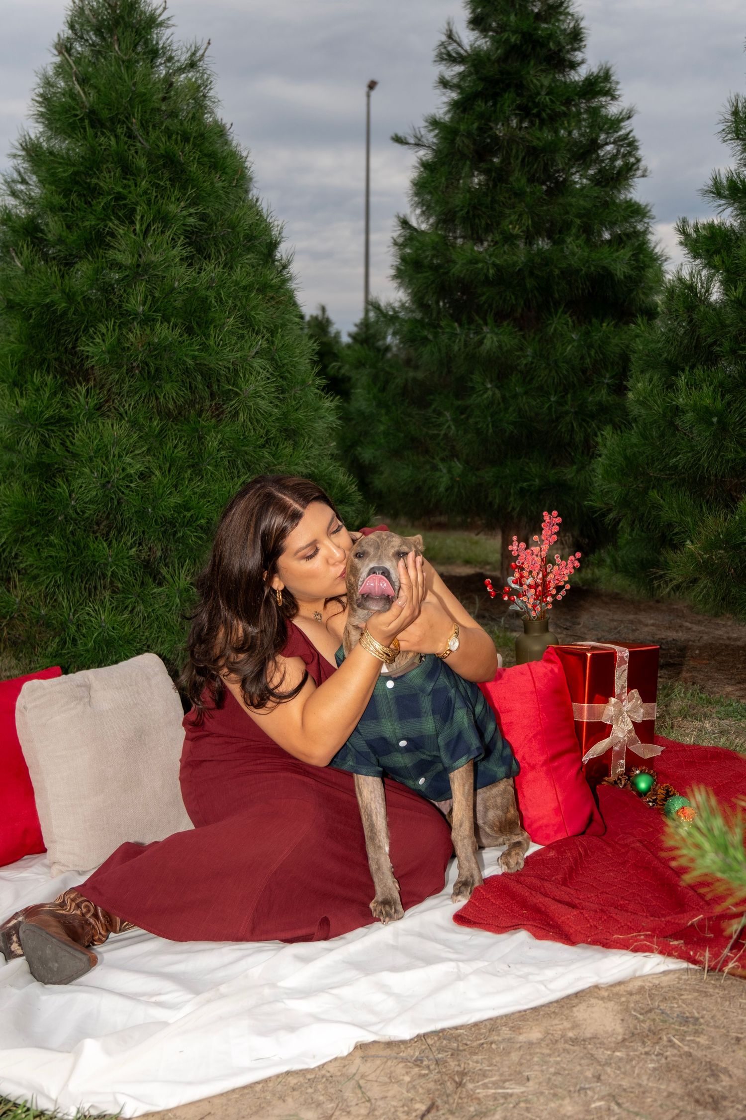 Woman in red dress kissing small dog in plaid shirt at Christmas tree farm with holiday decorations.