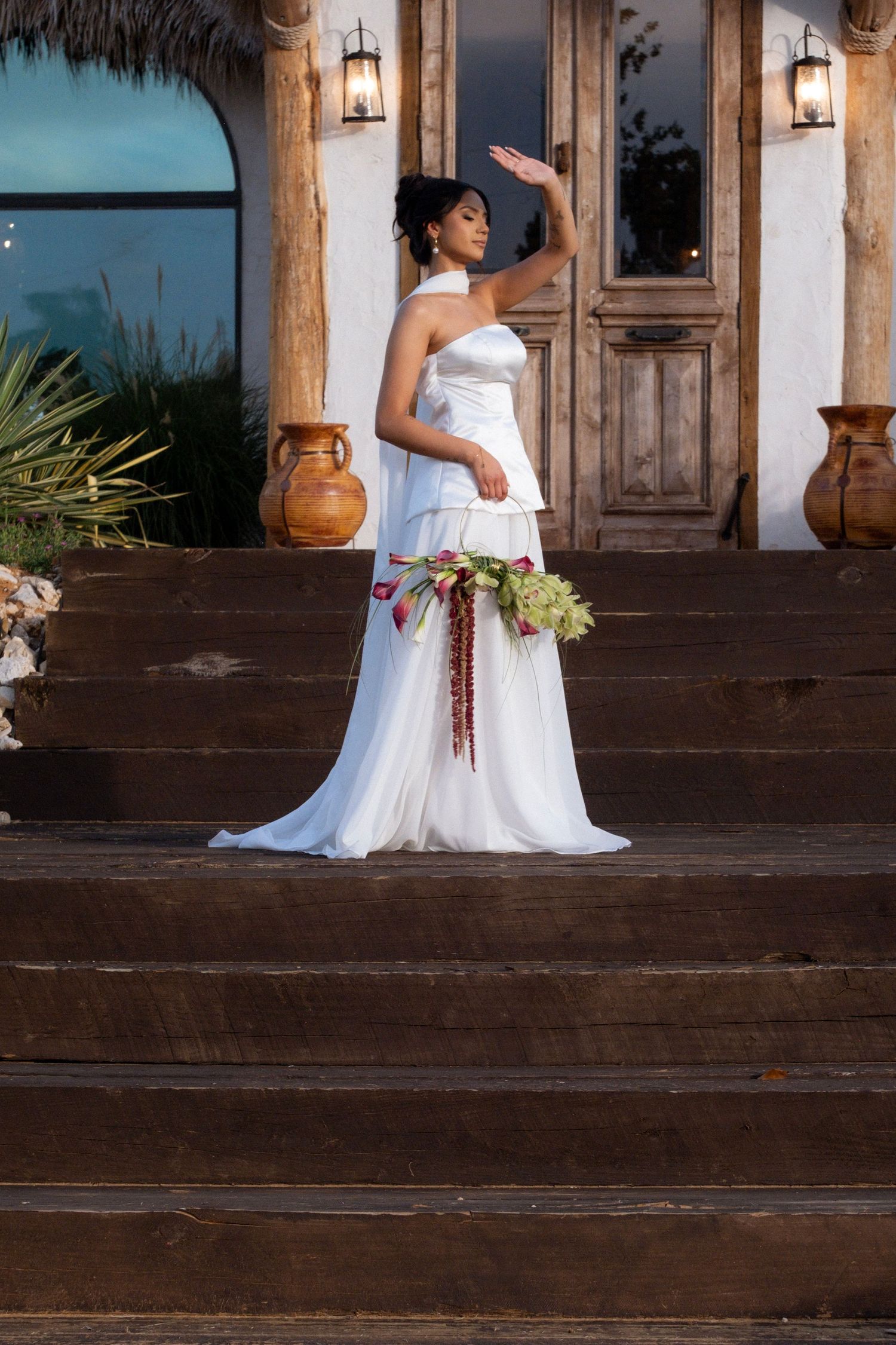 Elegant bride in white halter gown holding floral bouquet posing on rustic wooden steps at dusk.