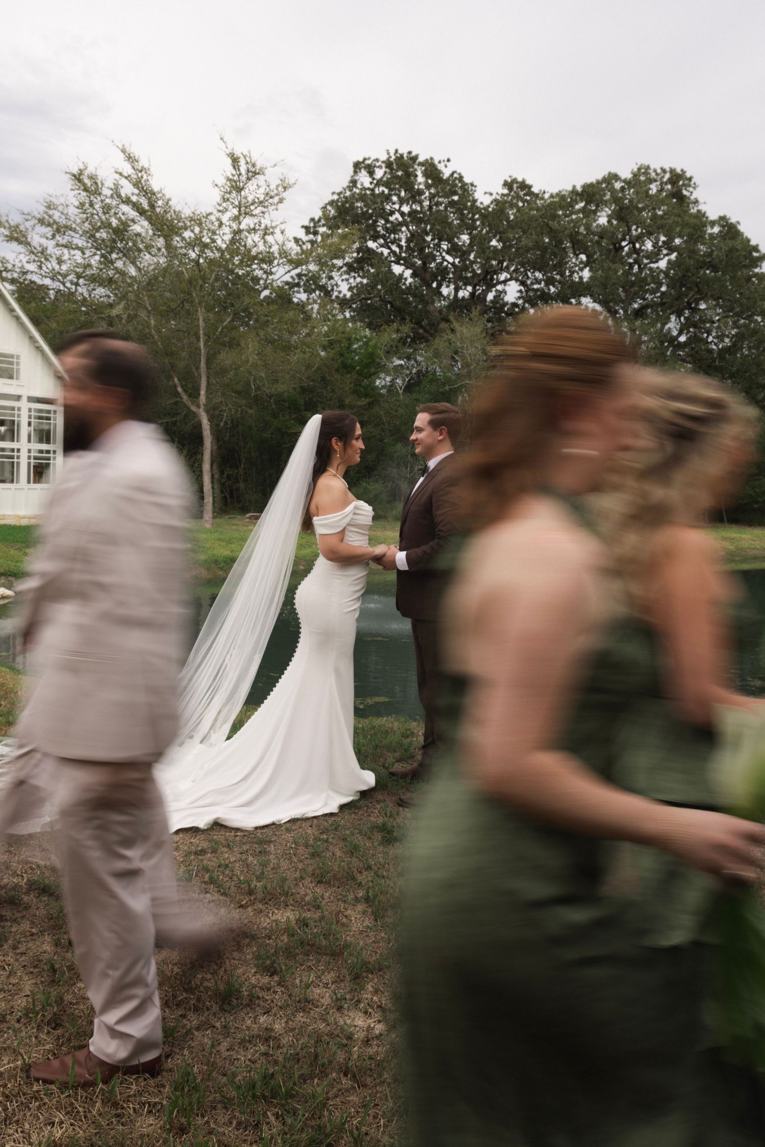 Bride in white gown and veil stands with groom outdoors as wedding guests in green and tan walk past.