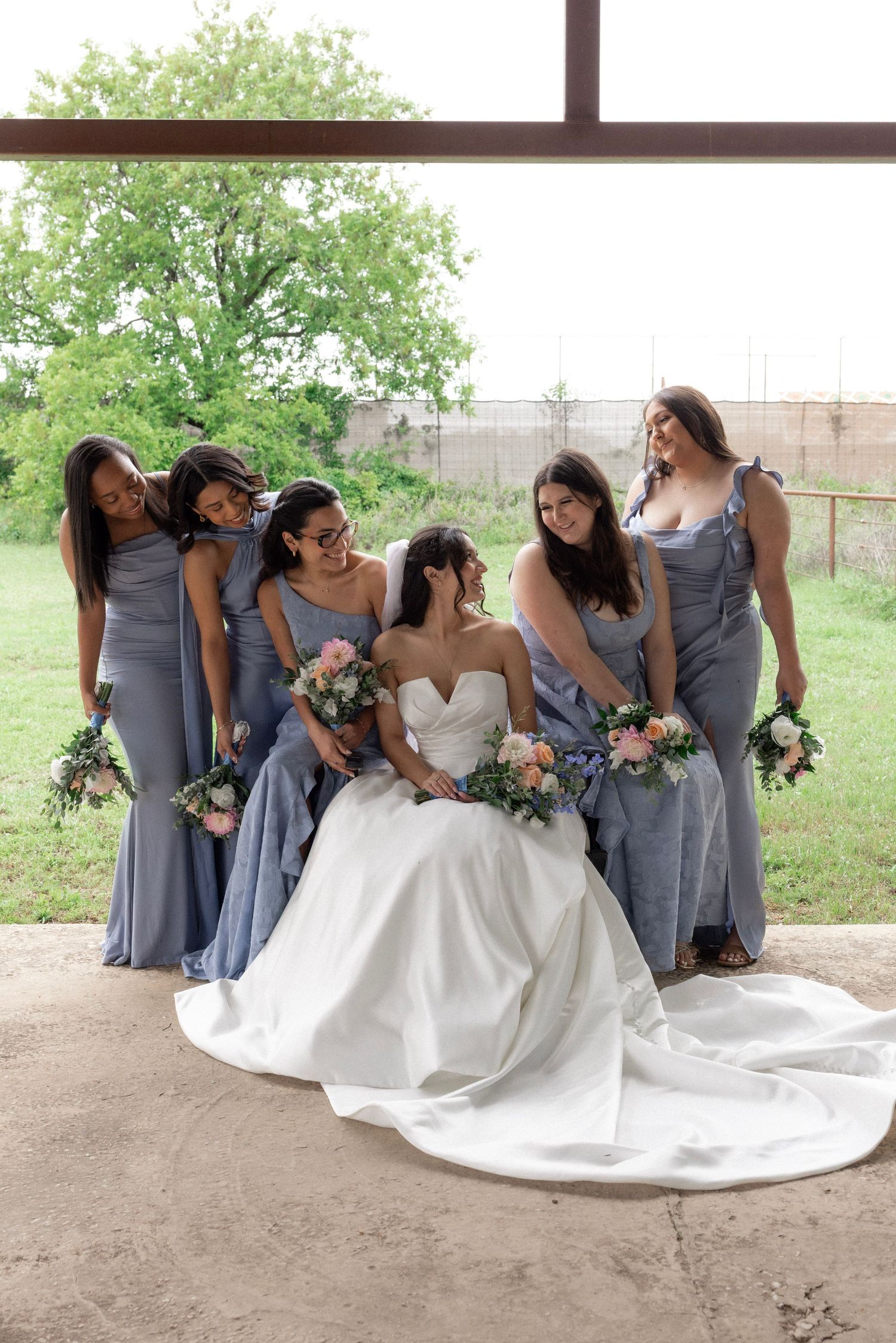 Bride seated in white ballgown surrounded by five bridesmaids in blue-gray gowns holding bouquets.
