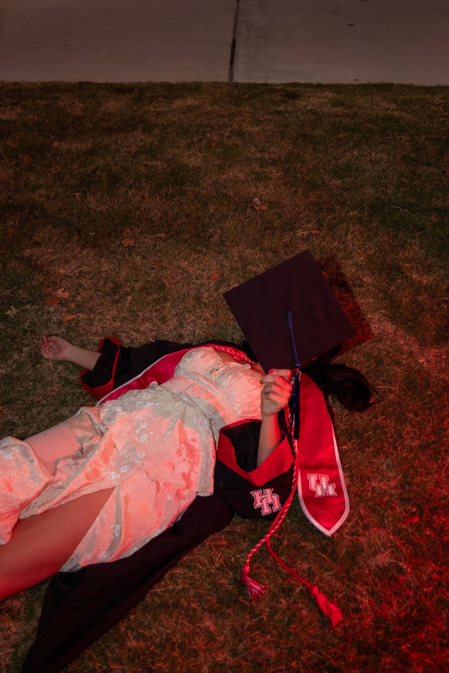 Graduate lying on grass at night with UH stole and cap covering face in red light.