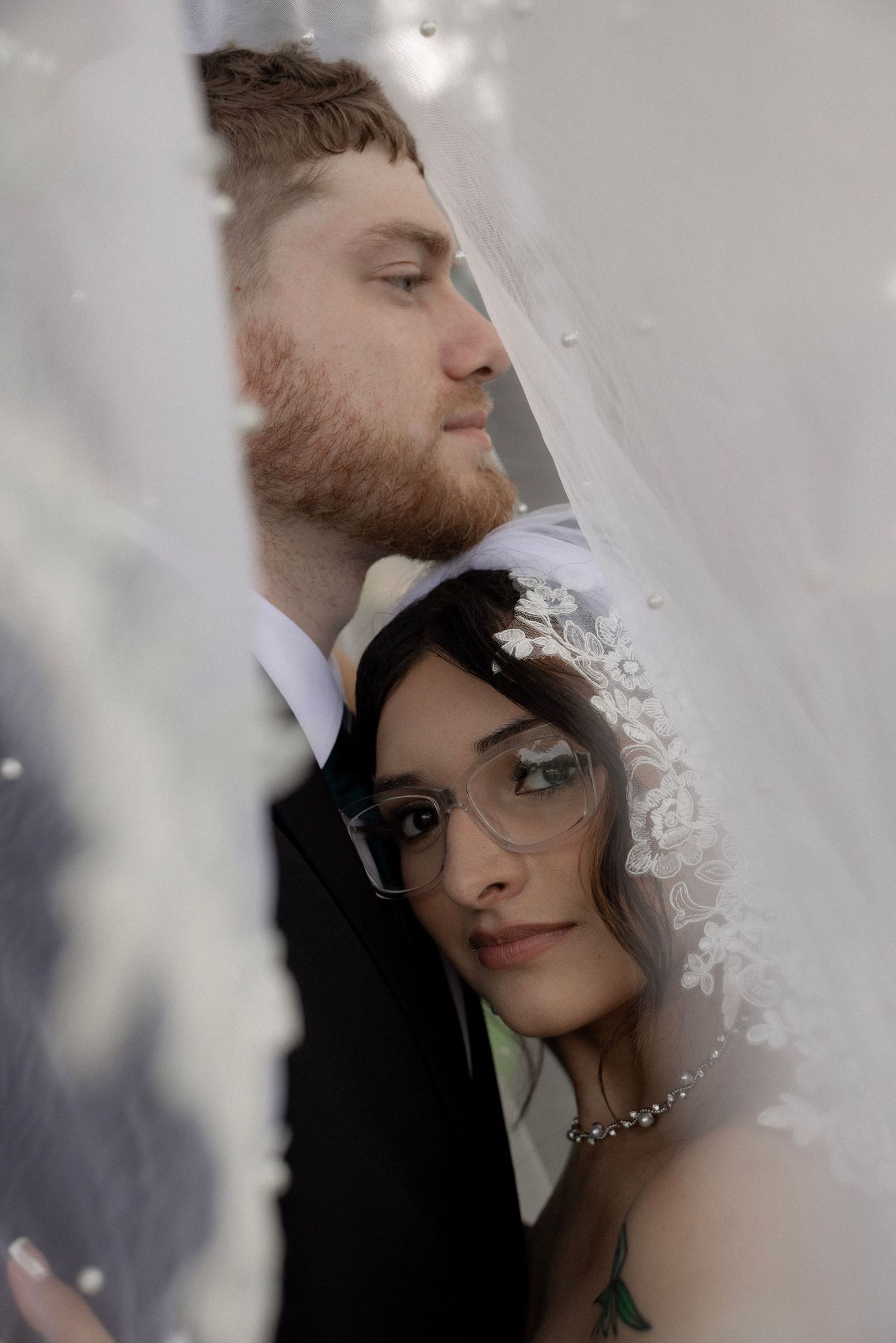 Intimate wedding portrait of bride with lace veil and glasses and groom with red beard seen through flowing white fabric.