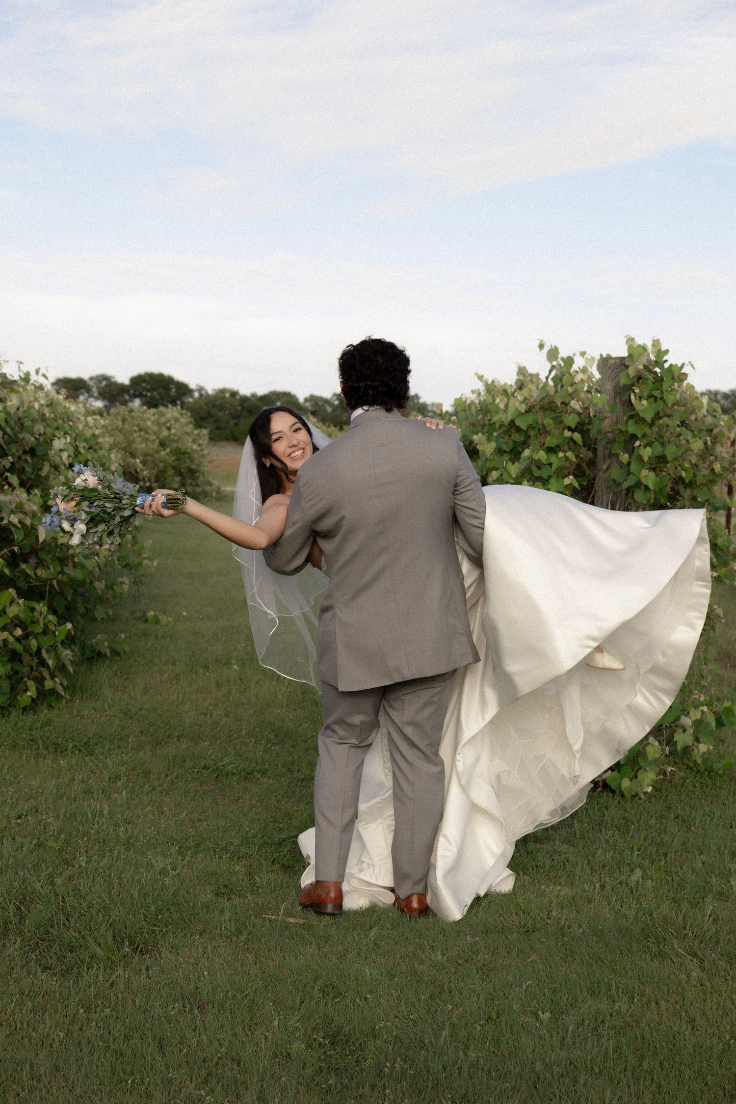Bride and groom walking through vineyard, bride smiling back as wedding dress flows in wind.