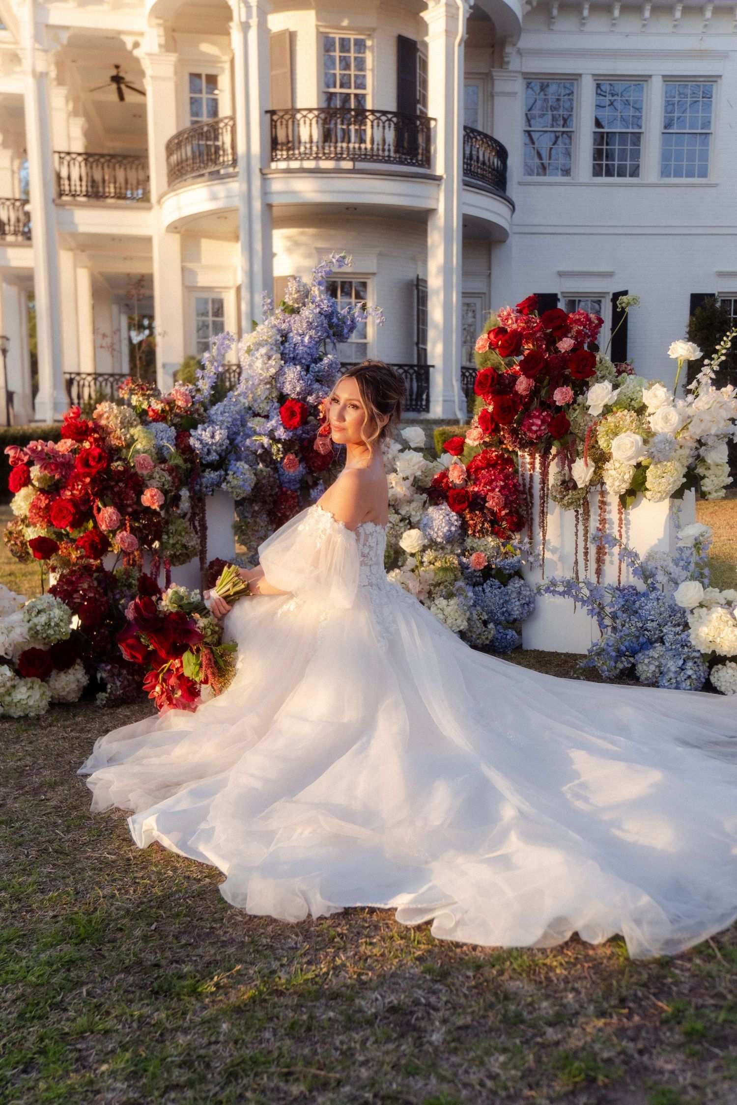 Bride in flowing white tulle ball gown poses among vibrant red and blue floral arrangements outdoors.