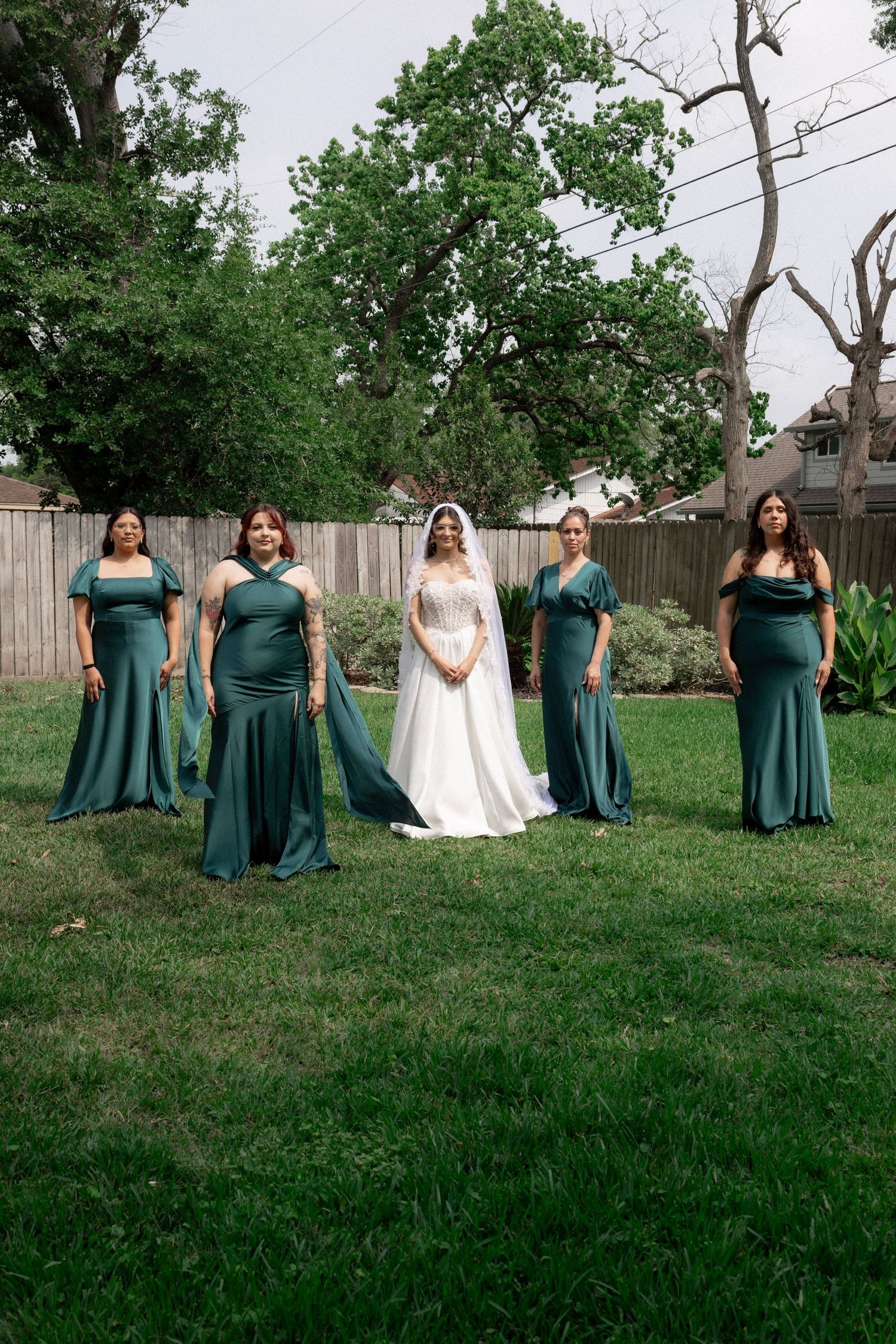 A bride in a white gown poses with four bridesmaids in emerald green dresses outdoors.