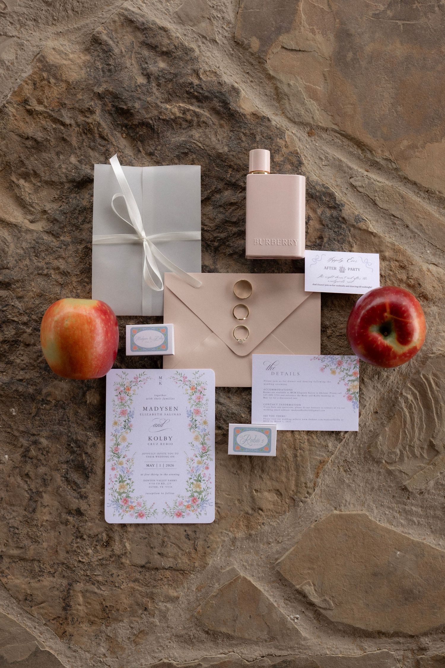Elegant wedding flat lay with floral invitations, rings, perfume bottle, and red apples on stone.