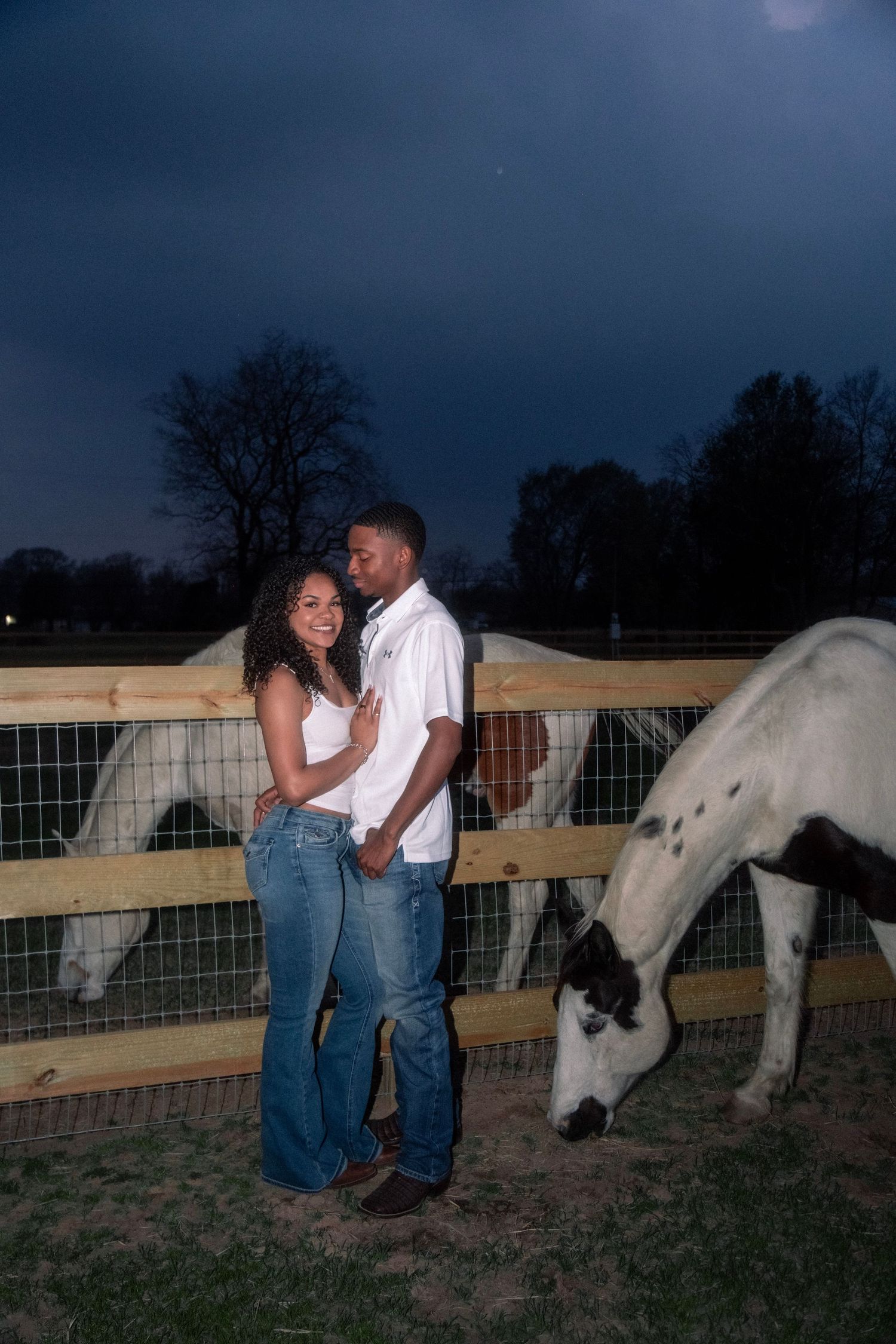 A couple embraces near a wooden fence at dusk while a white horse grazes beside them on a farm.