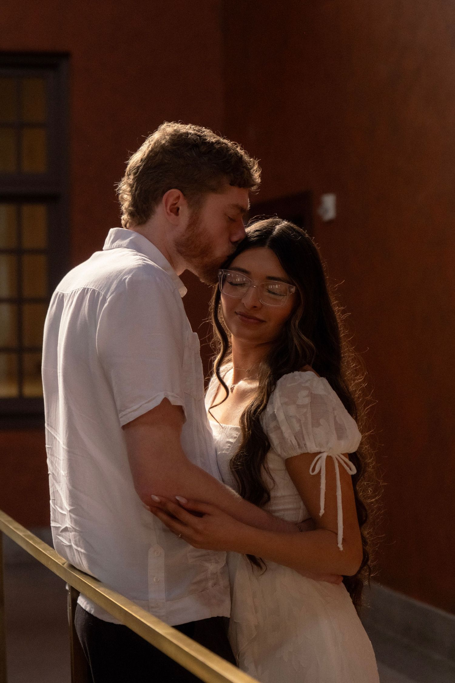 Couple shares tender moment as man kisses womans forehead near golden railing in warm sunlit hallway.