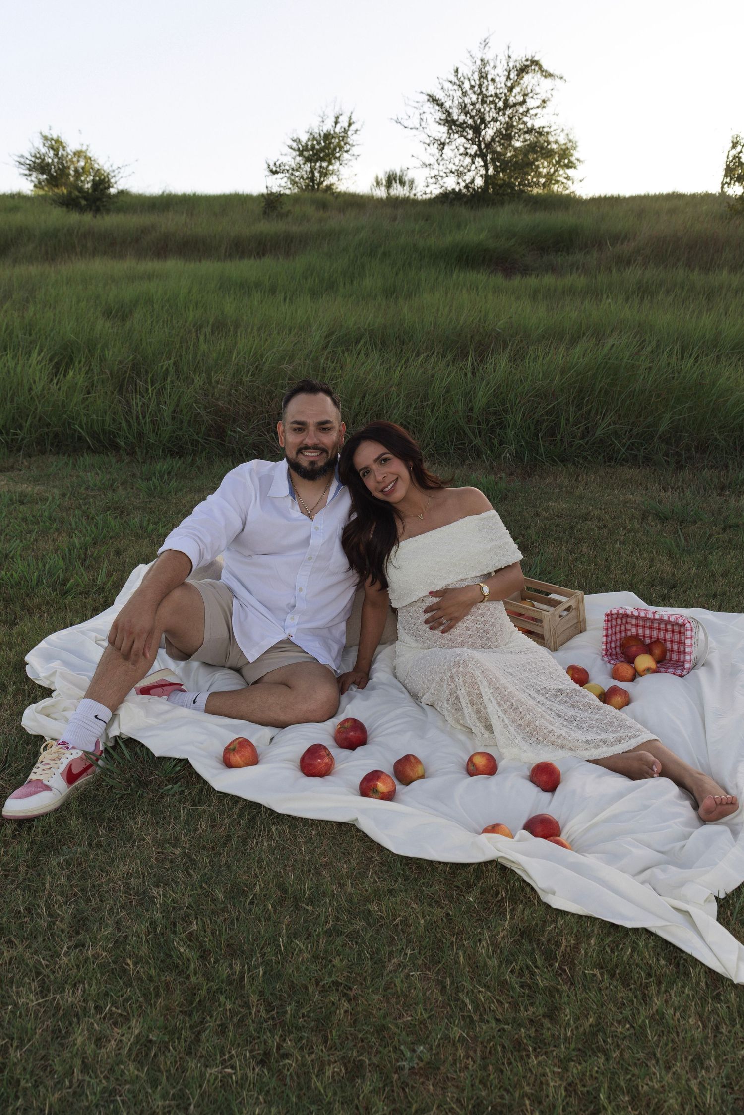 Couple sitting on white blanket outdoors for a romantic picnic with apples, surrounded by green grass at dusk.