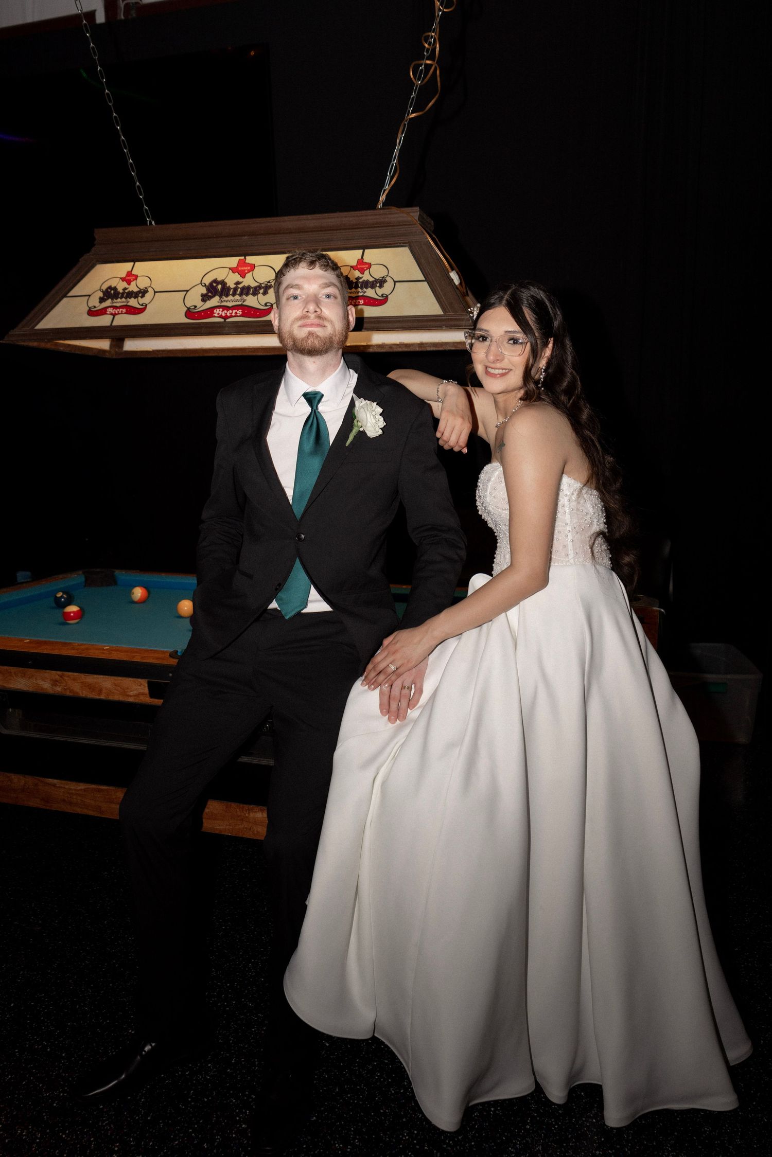 Bride and groom pose by pool table under Shiner beer light in fun wedding portrait.