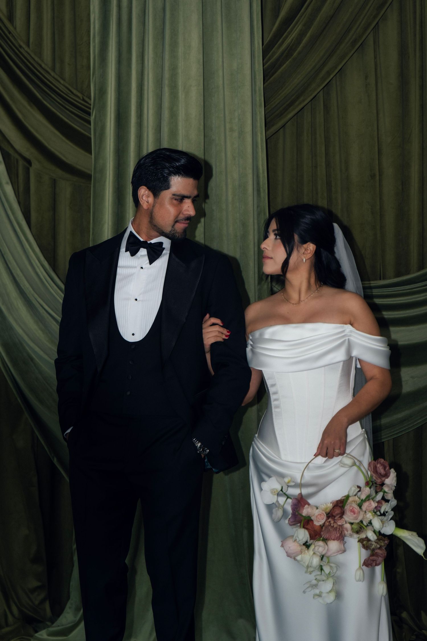 Bride and groom in black tuxedo stand elegantly before draped olive green velvet curtains, exchanging a loving glance.