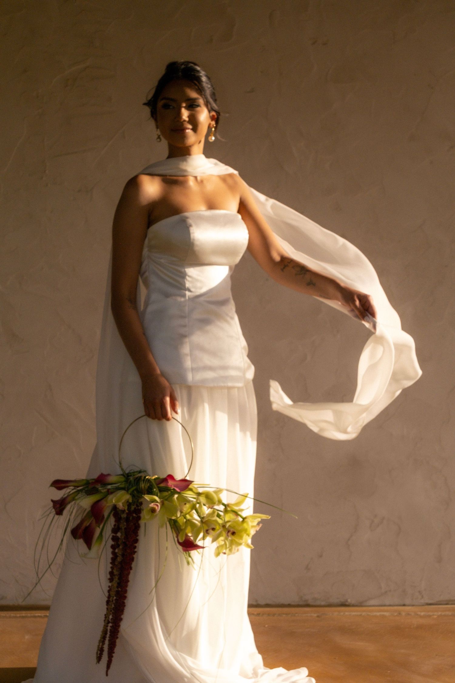 Bride in strapless white gown with flowing scarf holds floral basket in warm golden sunlight.