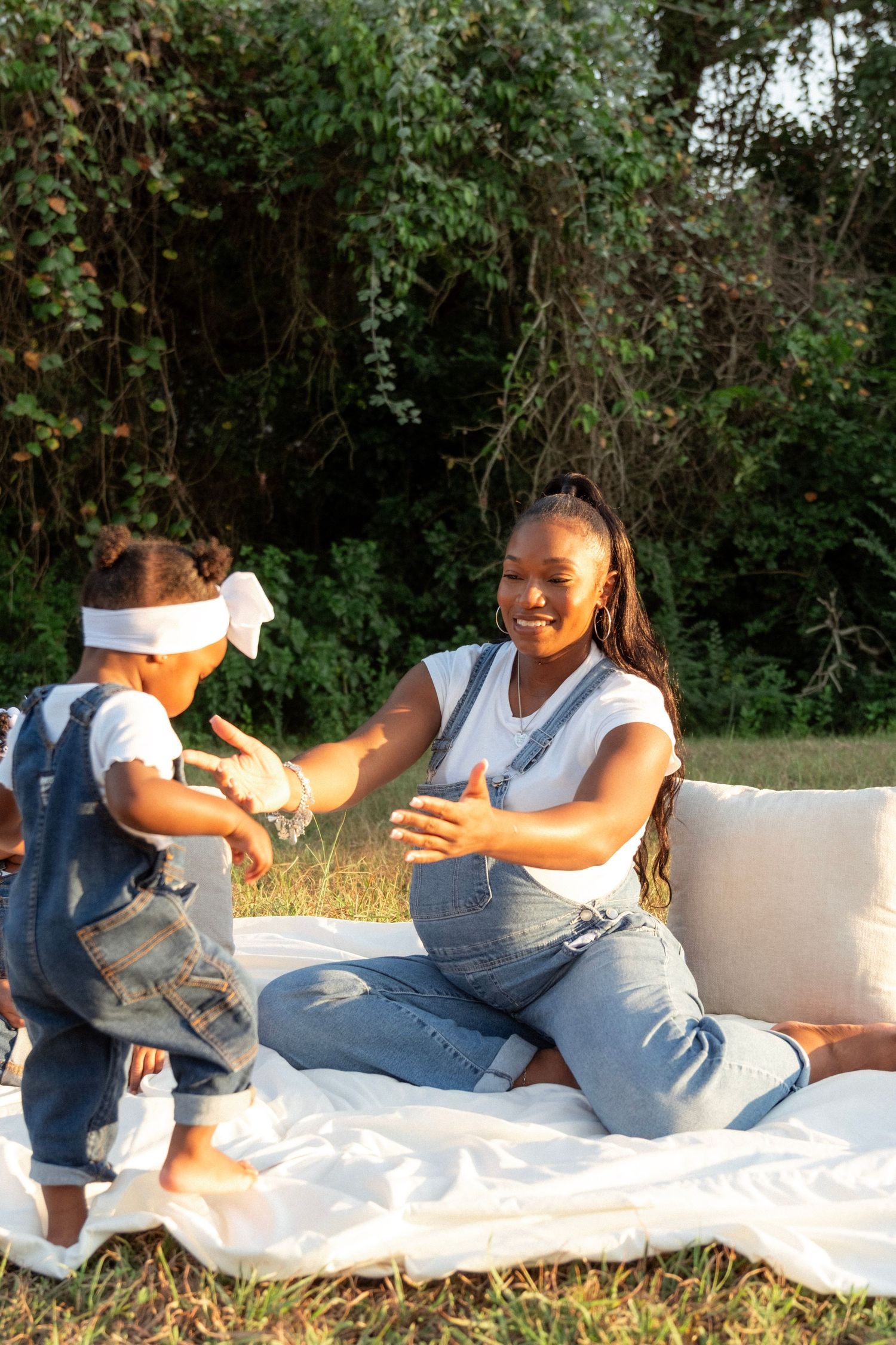 Pregnant woman in denim overalls playing with toddler on a white blanket in a sunny outdoor maternity session.