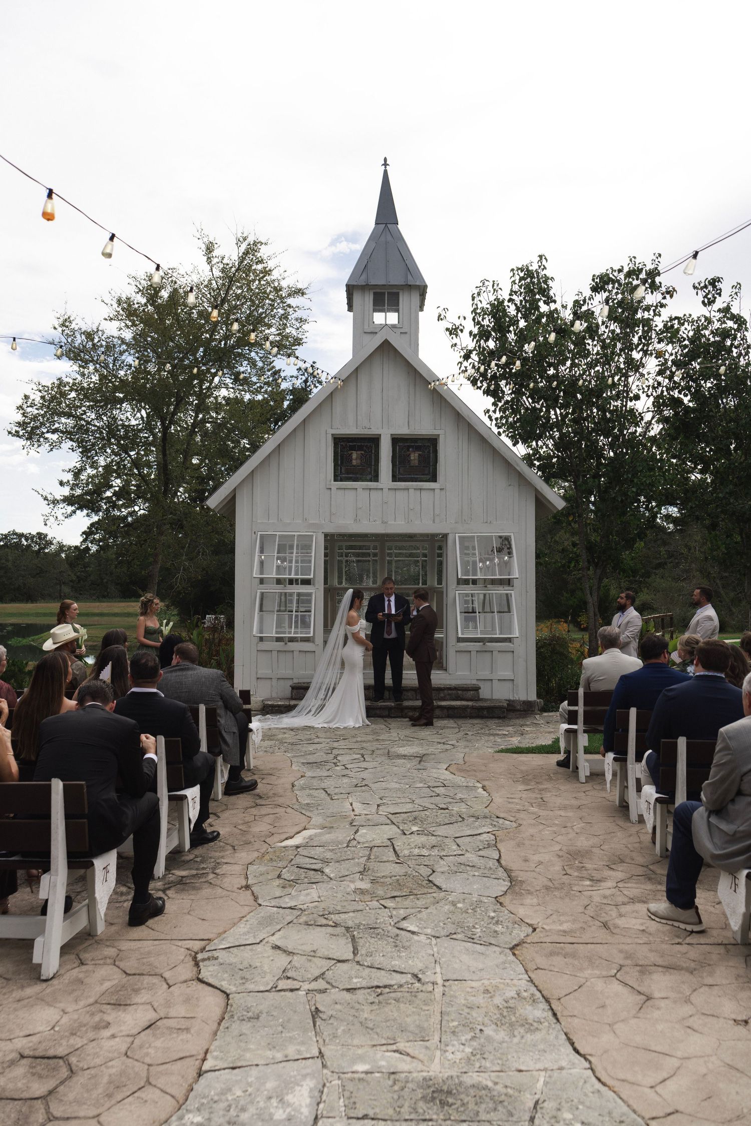 Outdoor wedding ceremony at a rustic white chapel with stone aisle, wooden pews, and string lights.